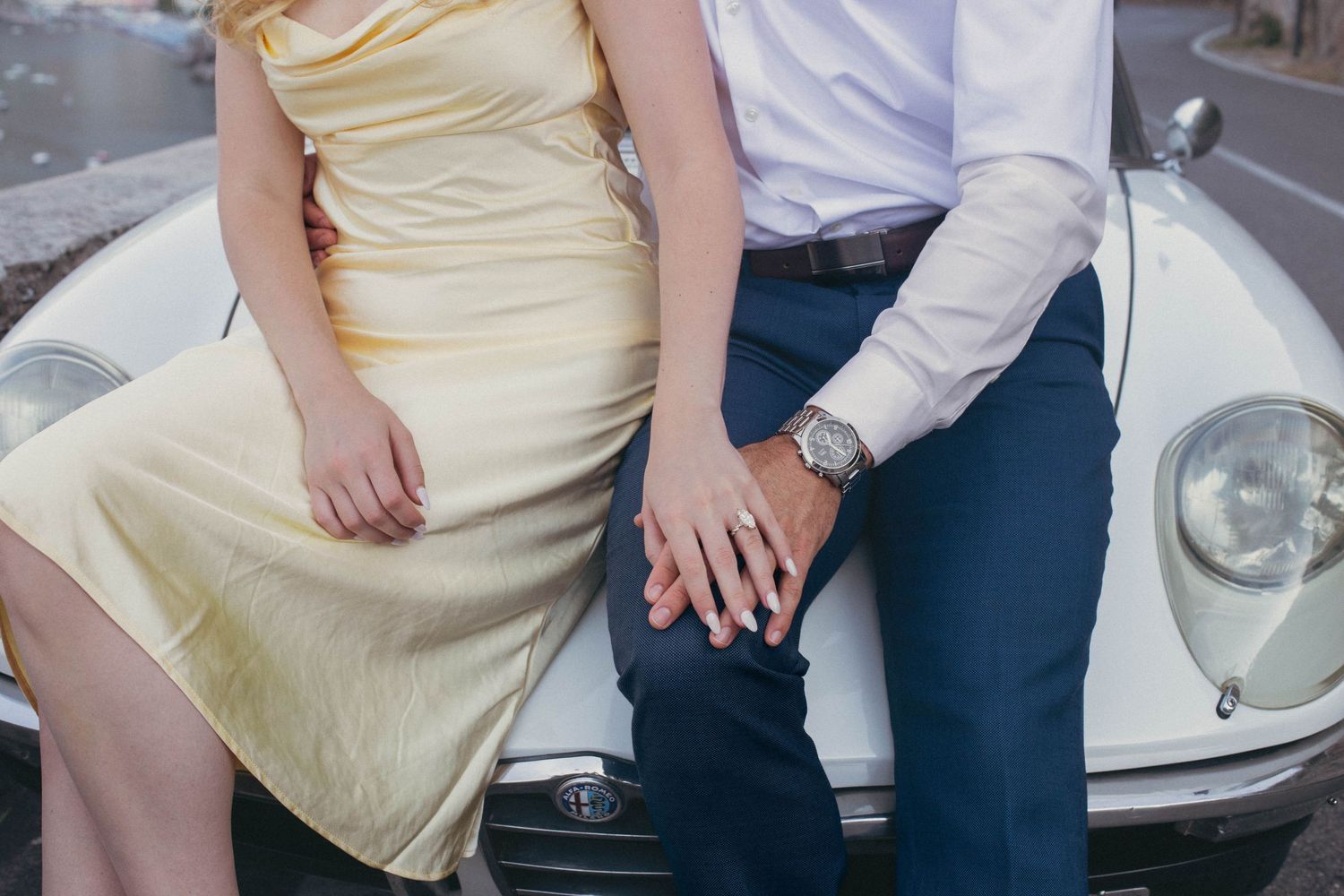 Couple in elegant attire sitting on a vintage white car, holding hands, wearing a watch and yellow dress.