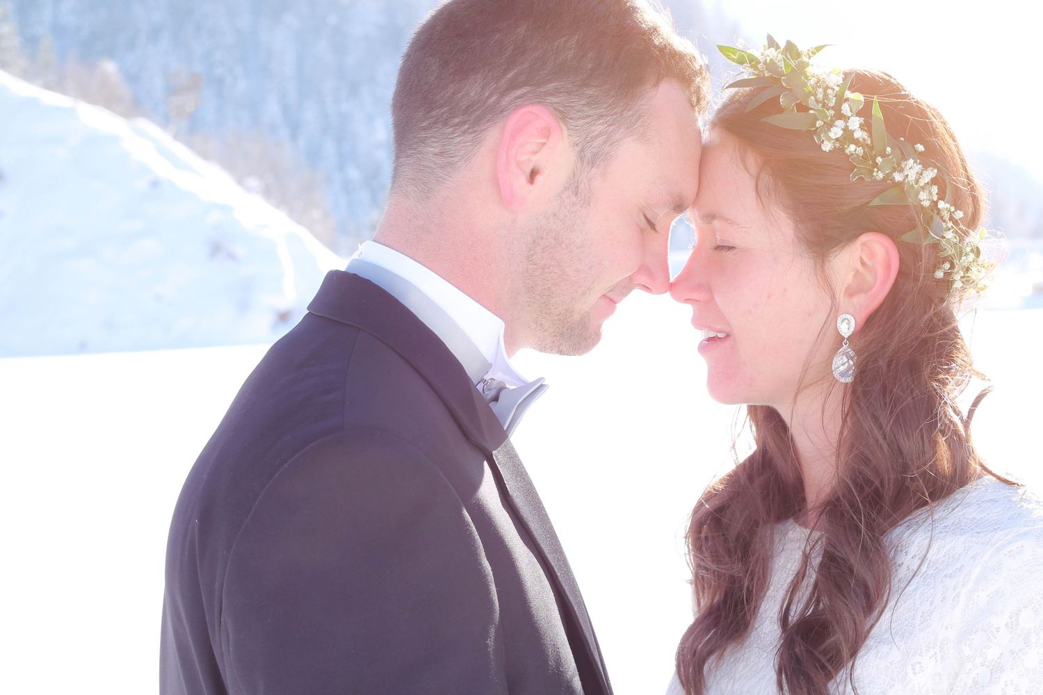 Romantic winter wedding couple touching foreheads in snowy landscape, bride wearing floral crown and groom in dark suit.
