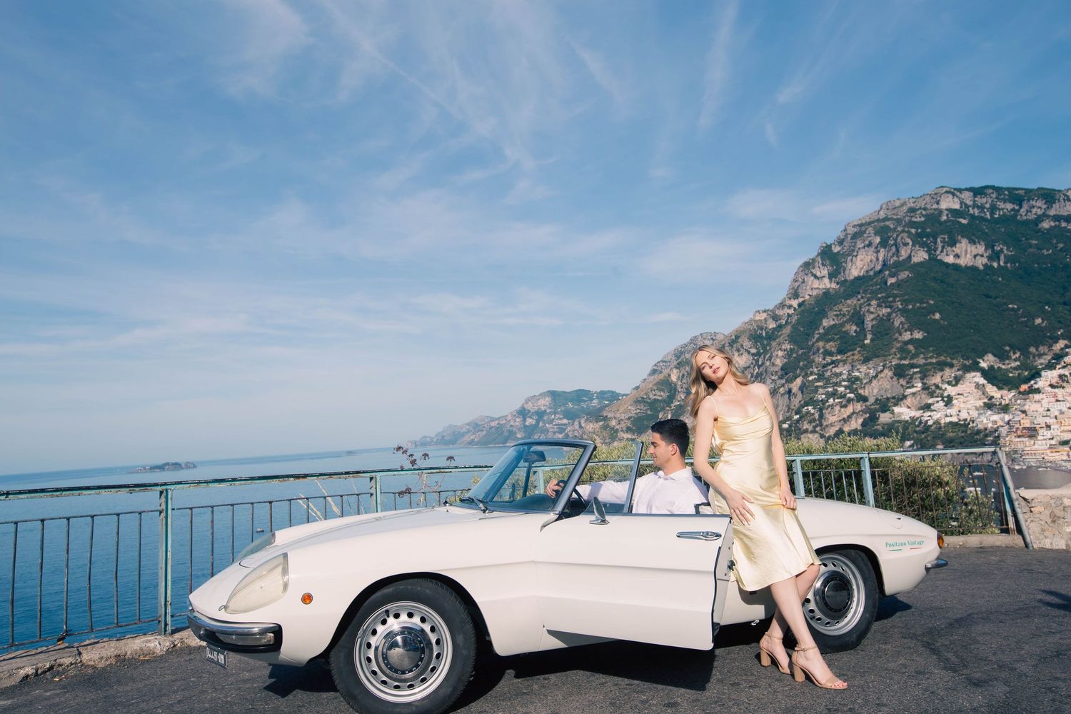 Woman in gold dress leaning on a classic white convertible with a man seated inside on the Amalfi Coast.