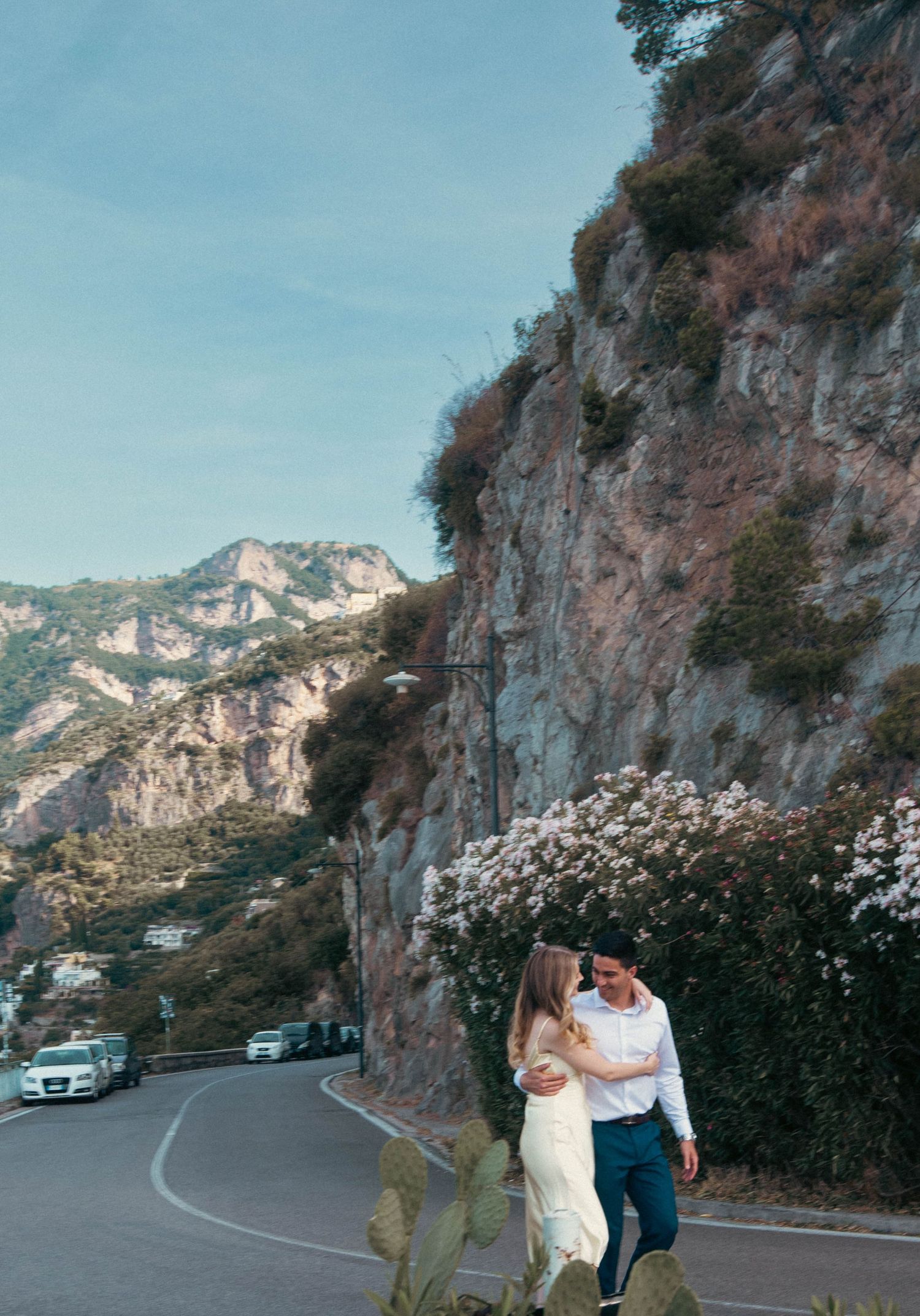 Couple embracing while walking along a winding Amalfi Coast road with dramatic cliffs and blooming flowers.