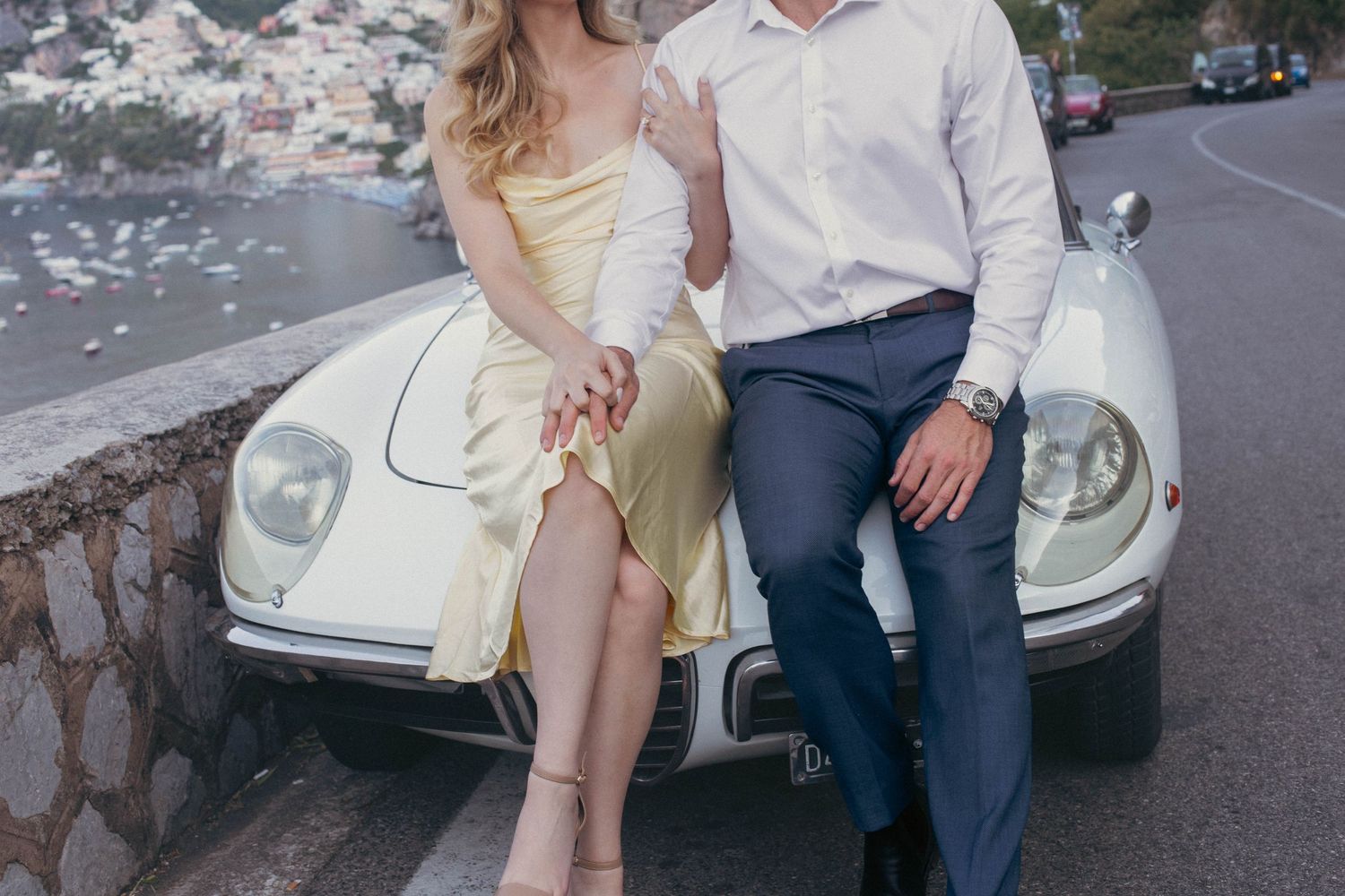 Couple posing on a vintage white sports car on a scenic Amalfi Coast road, woman in yellow dress, man in white shirt.