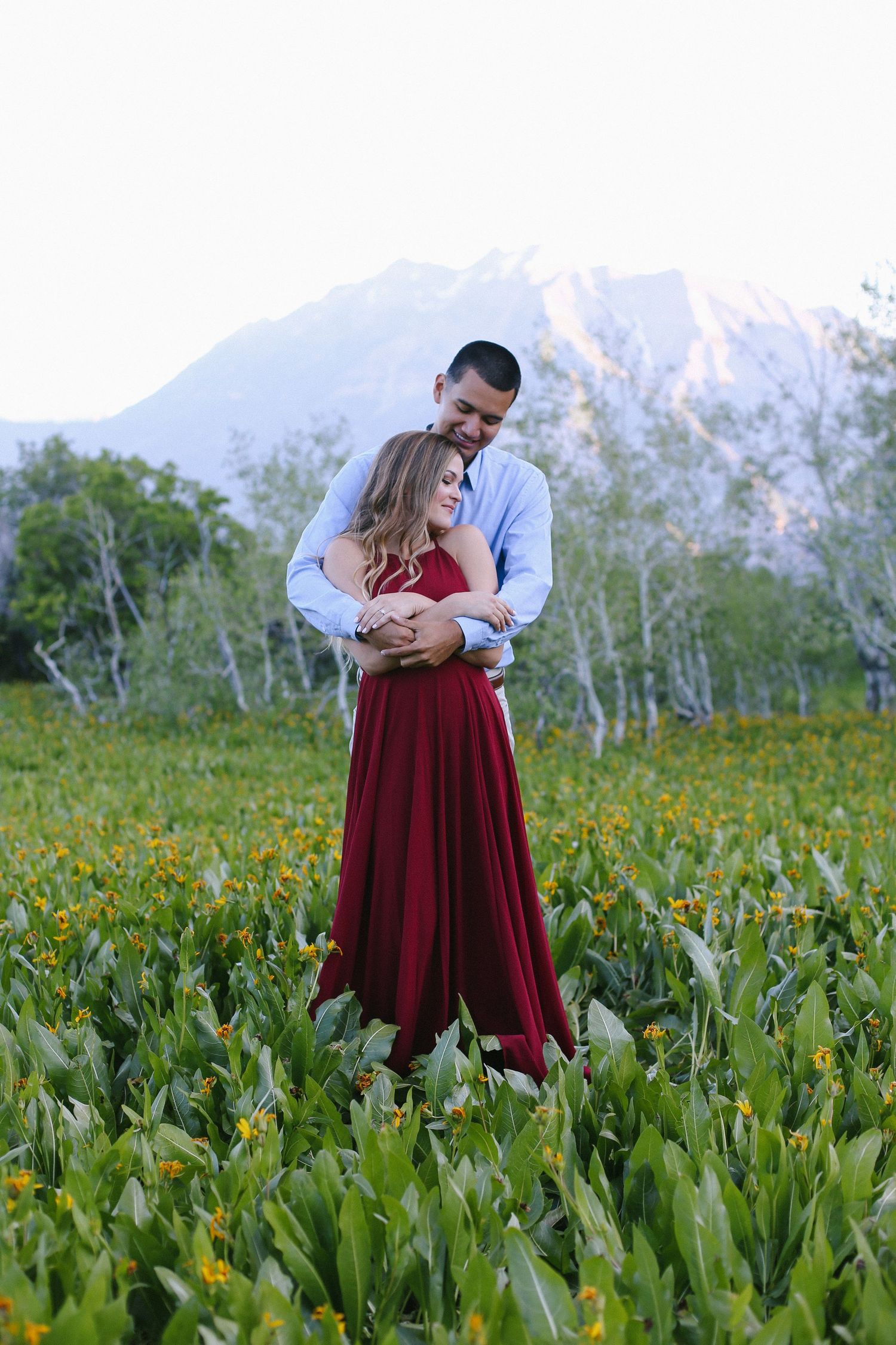 Couple embracing in a lush green meadow with yellow wildflowers and a snow-capped mountain backdrop.