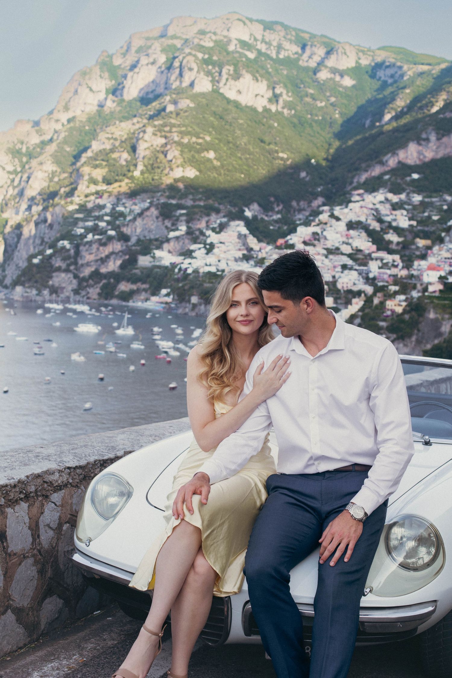 Romantic couple leaning against a vintage white car with Positano cliffside village and blue sea in background.