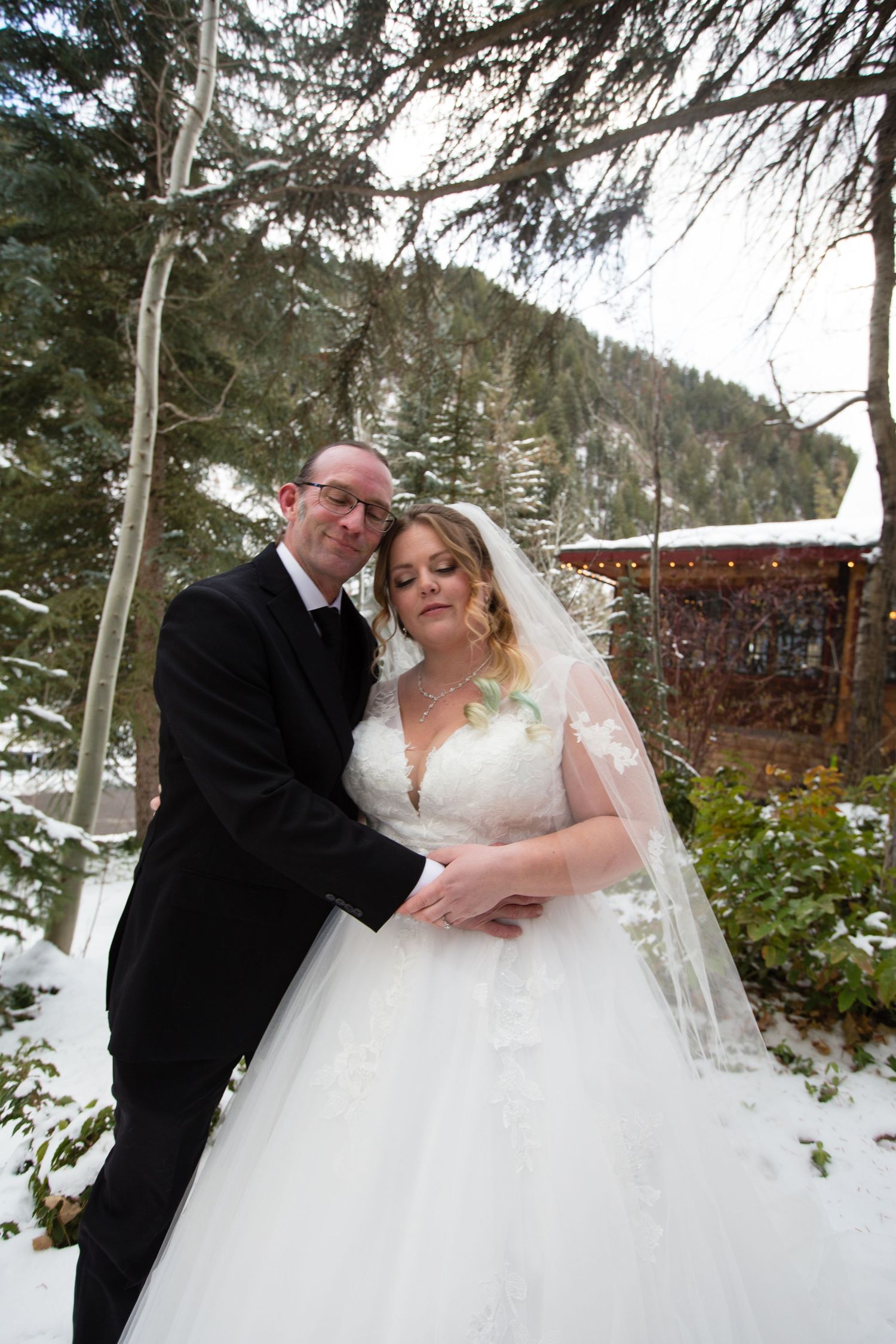 Newlyweds share an intimate moment in a snow-covered mountain setting with evergreen trees.