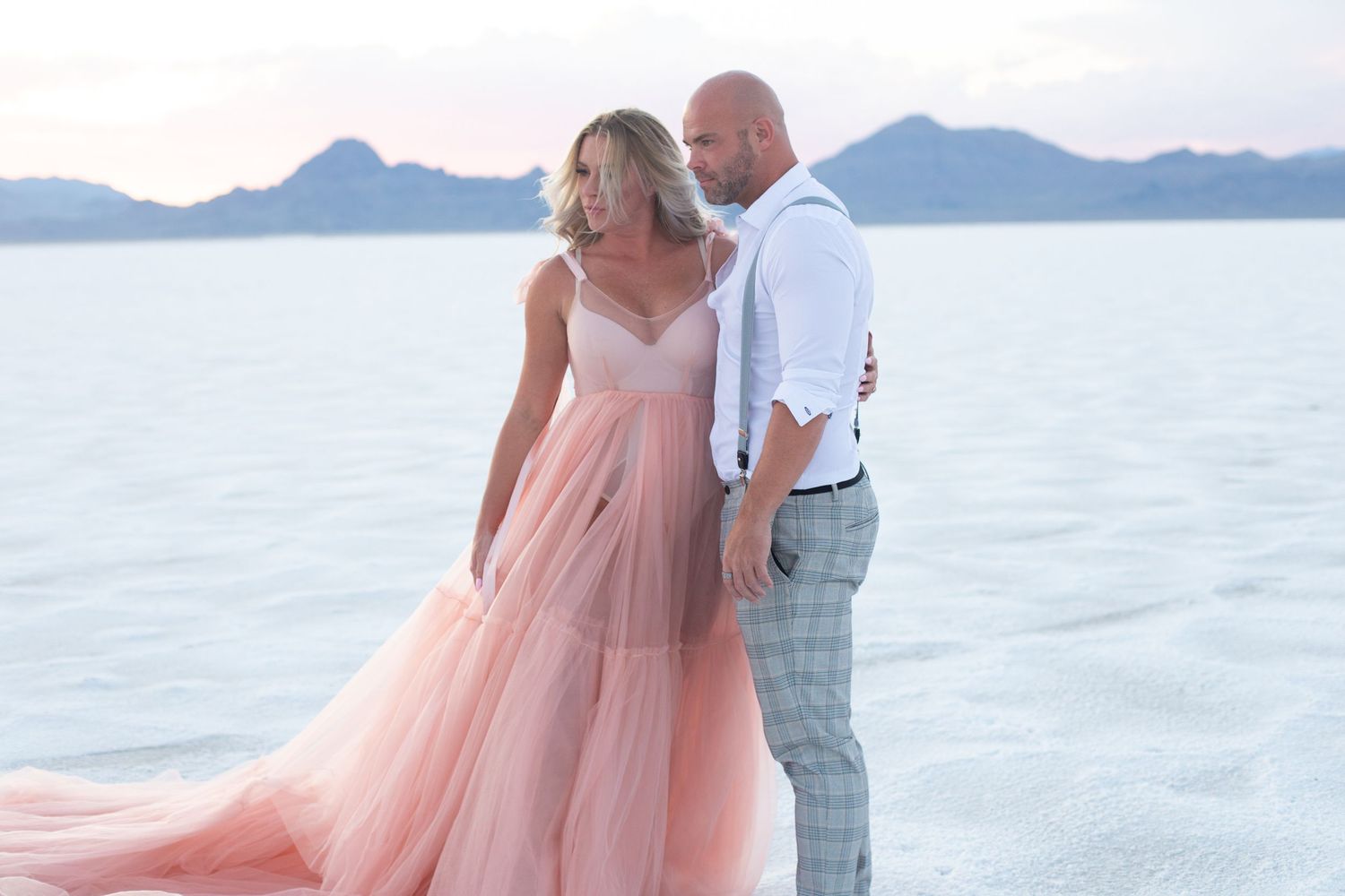 Couple poses on salt flats at sunset with mountains in background.