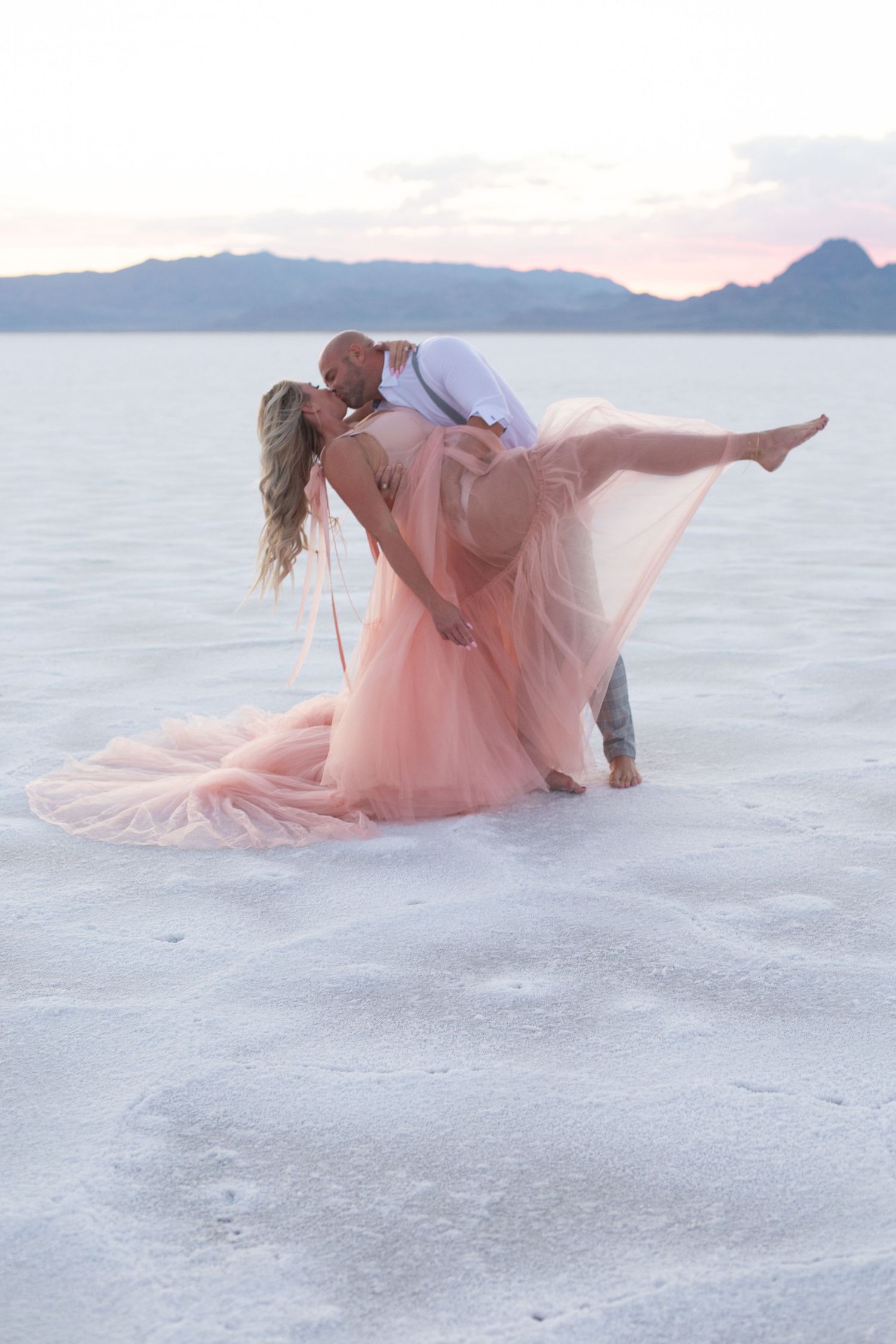 Dramatic dip pose with flowing pink dress on white salt flats at sunset.