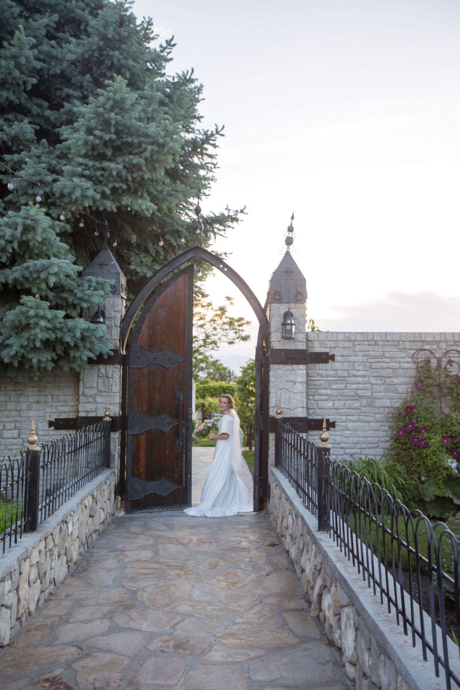 A stone pathway with wrought iron railings leads to an arched wooden door surrounded by evergreen trees.