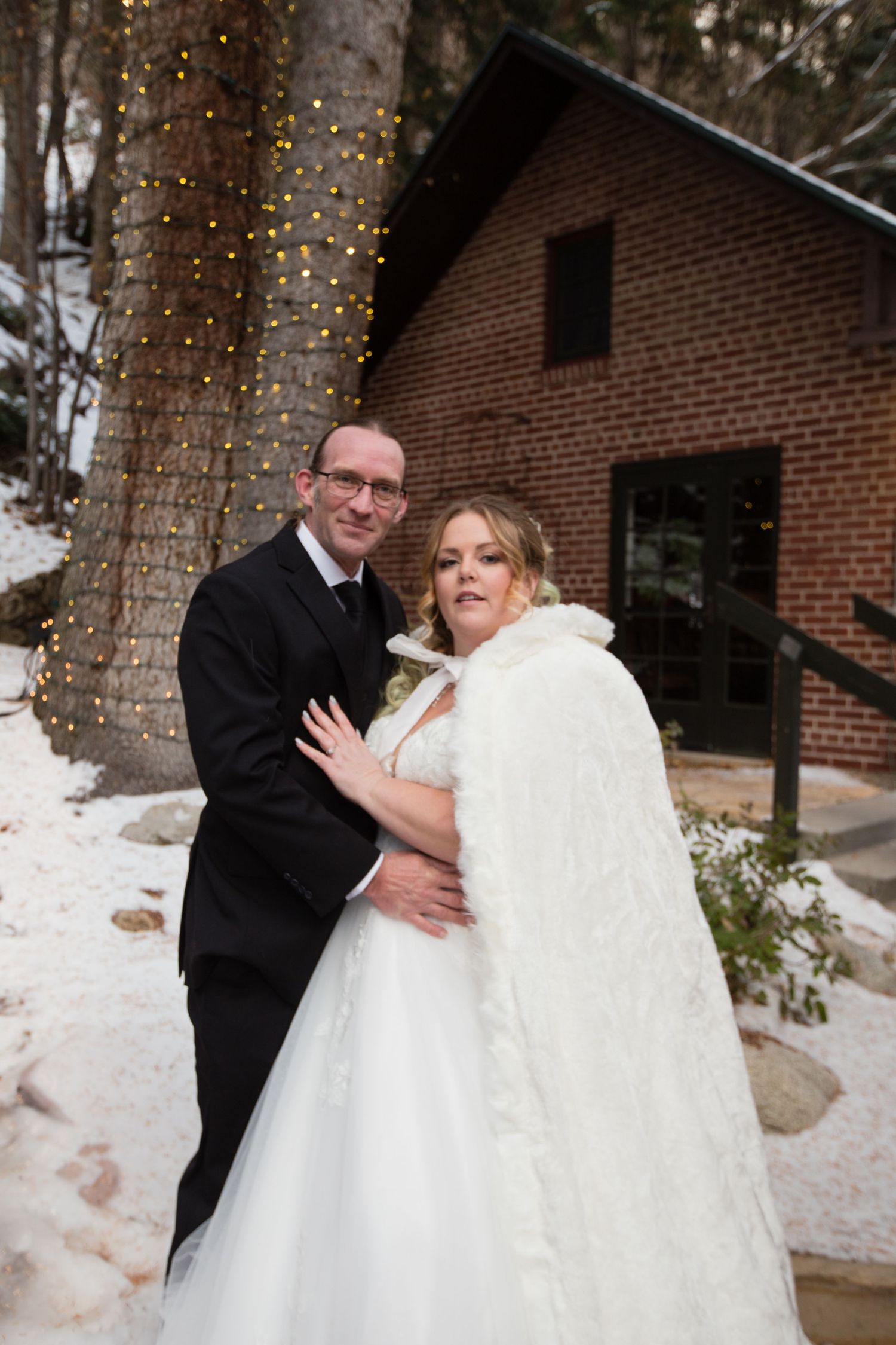 A winter wedding photo featuring a rustic brick cabin with twinkling lights wrapped around nearby trees.