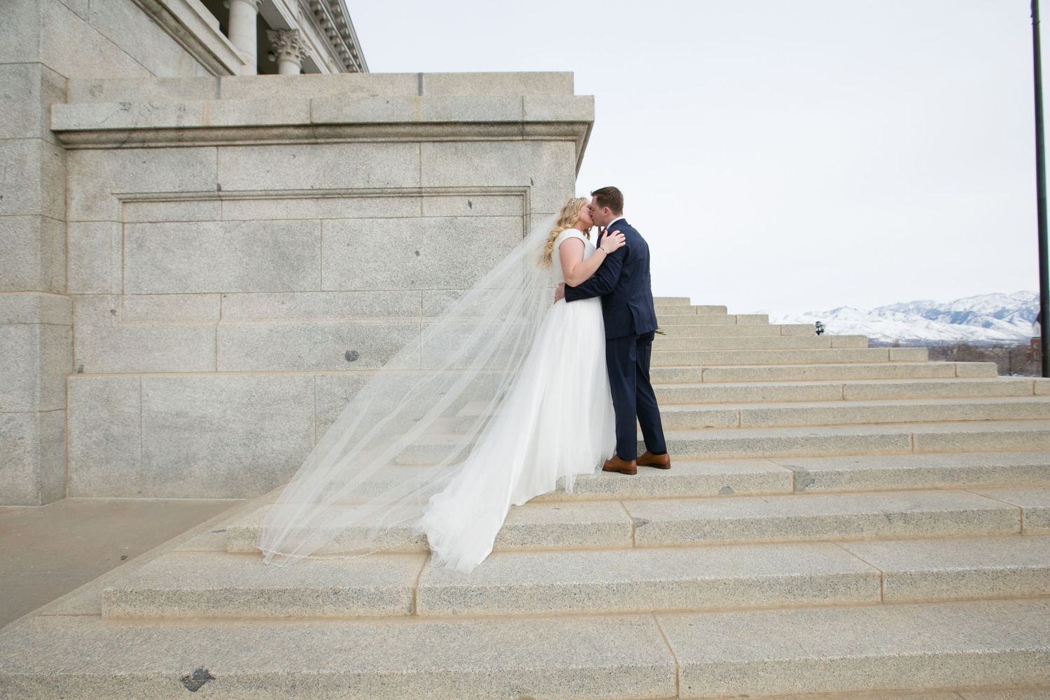 Newlyweds embracing on grand stone steps with flowing white veil.
