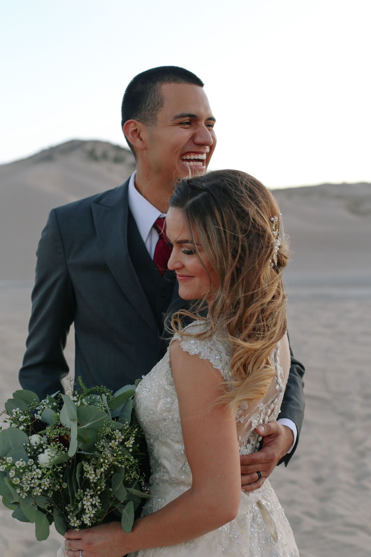 Joyful couple sharing intimate moment on sandy dunes with bouquet of greenery.
