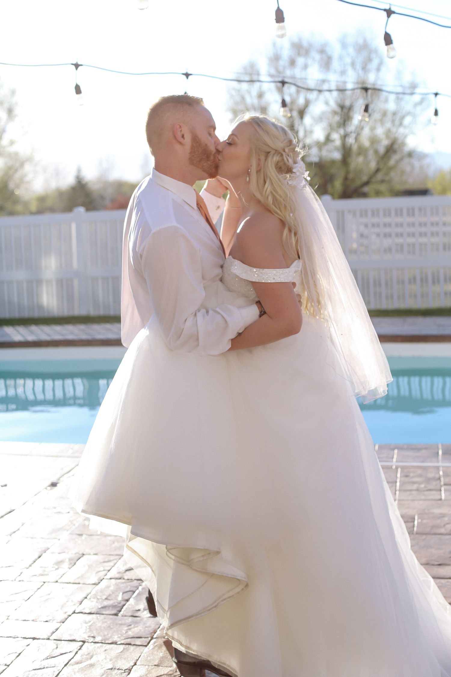 Couple in wedding attire dancing and embracing by a swimming pool on a sunny day.