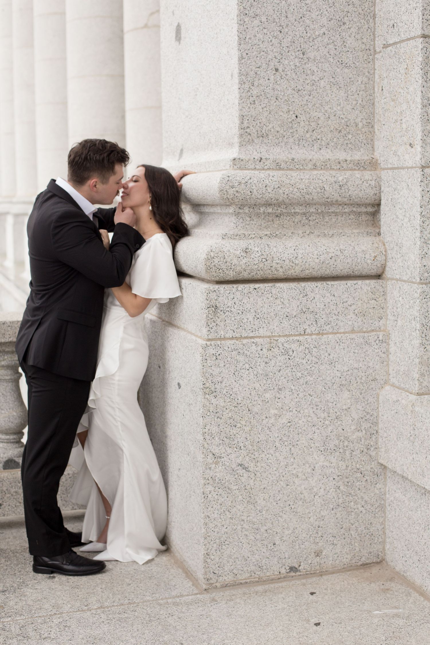 A couple shares a romantic moment between white stone columns at a grand building.