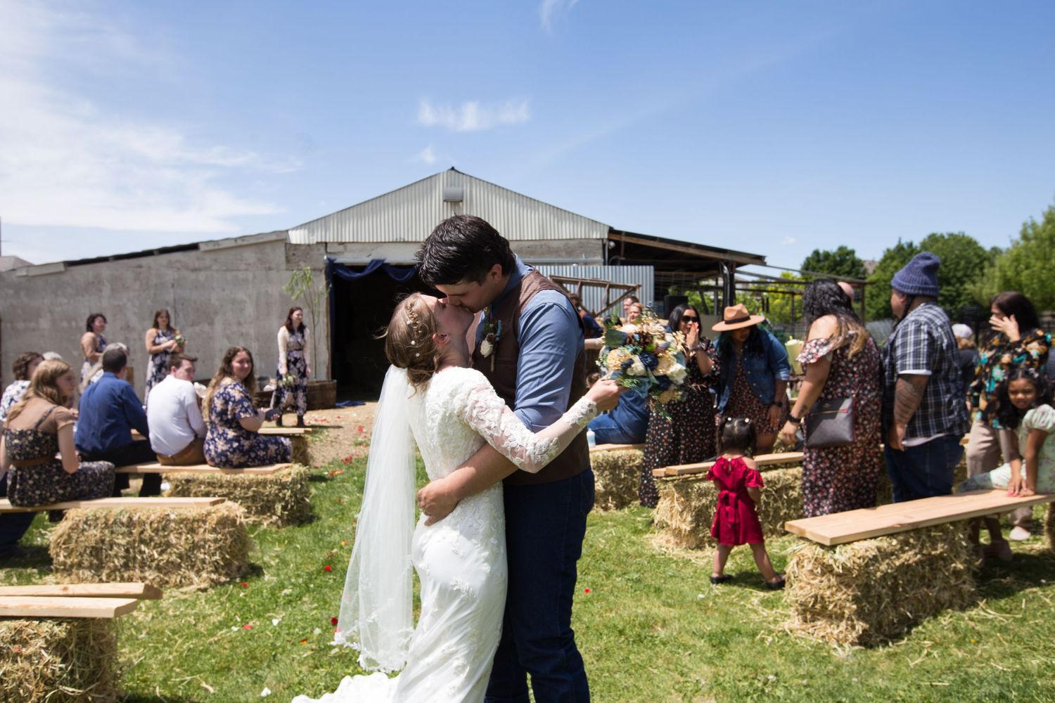 Bride and groom share first kiss surrounded by guests seated on hay bales at outdoor farm wedding.