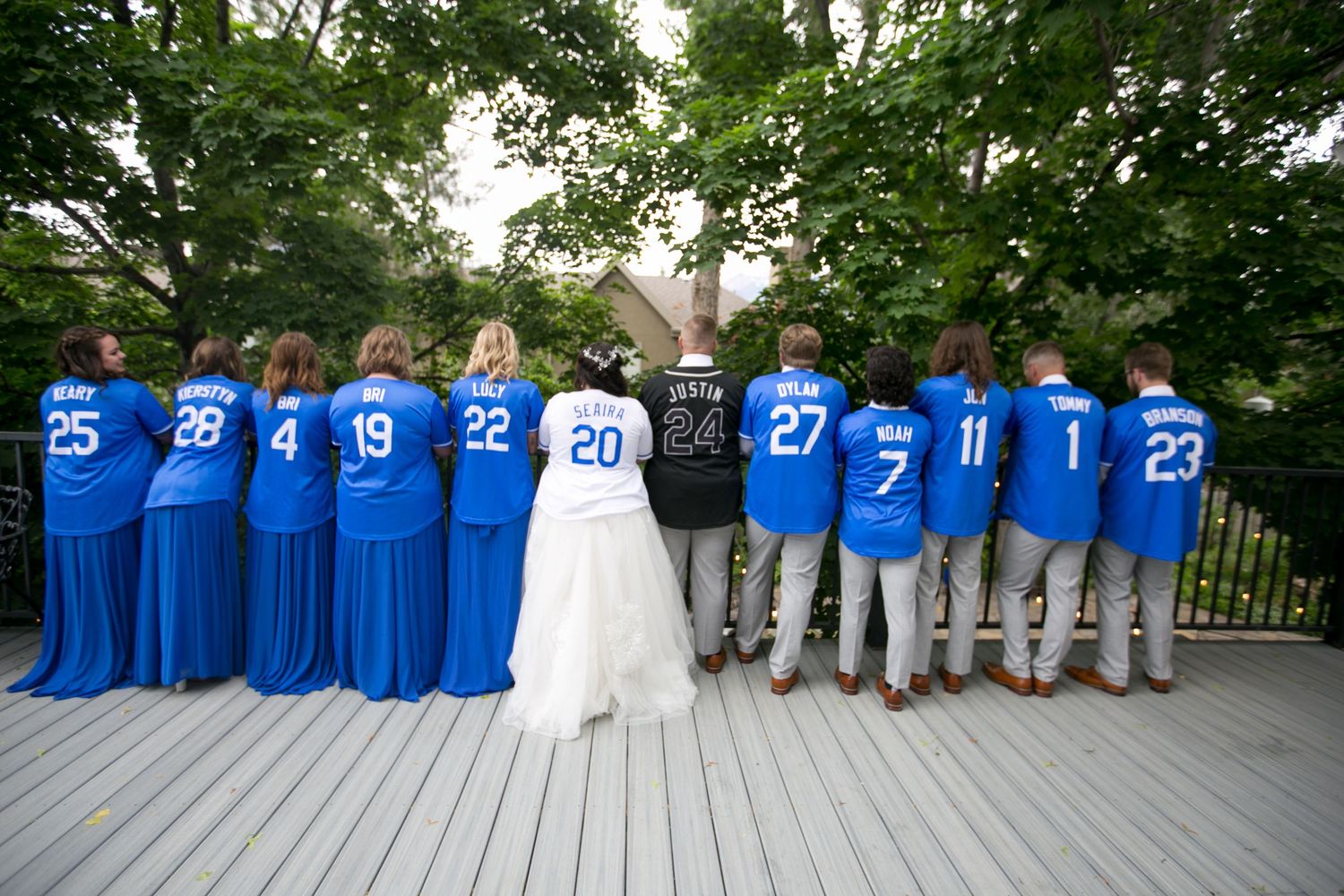 Wedding party in blue sports jerseys numbered 1-28 poses on wooden deck, bride and groom centered in white.