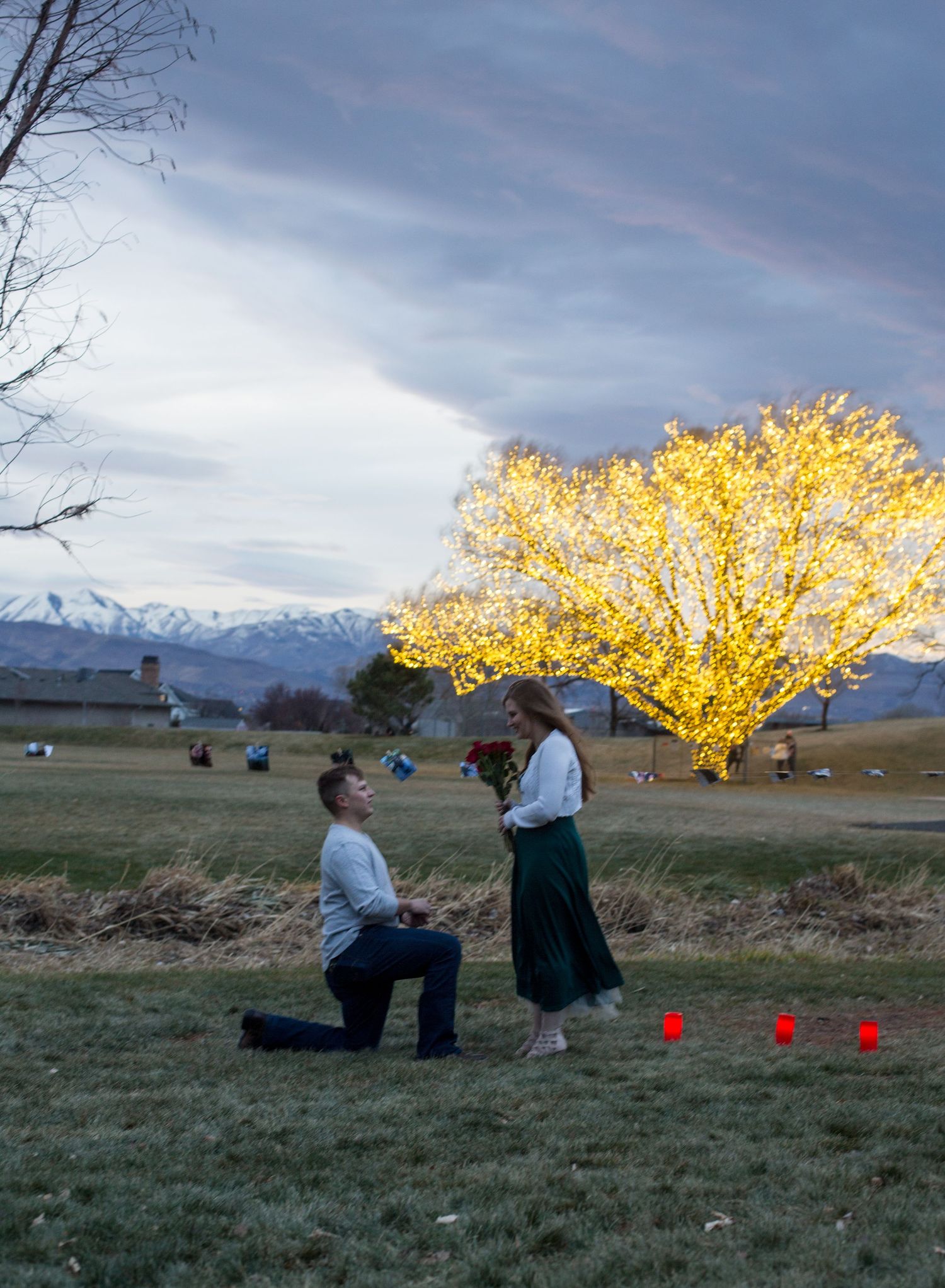 Man proposes on one knee to woman at dusk, with a glowing tree, red candles, and snowy mountains in background.
