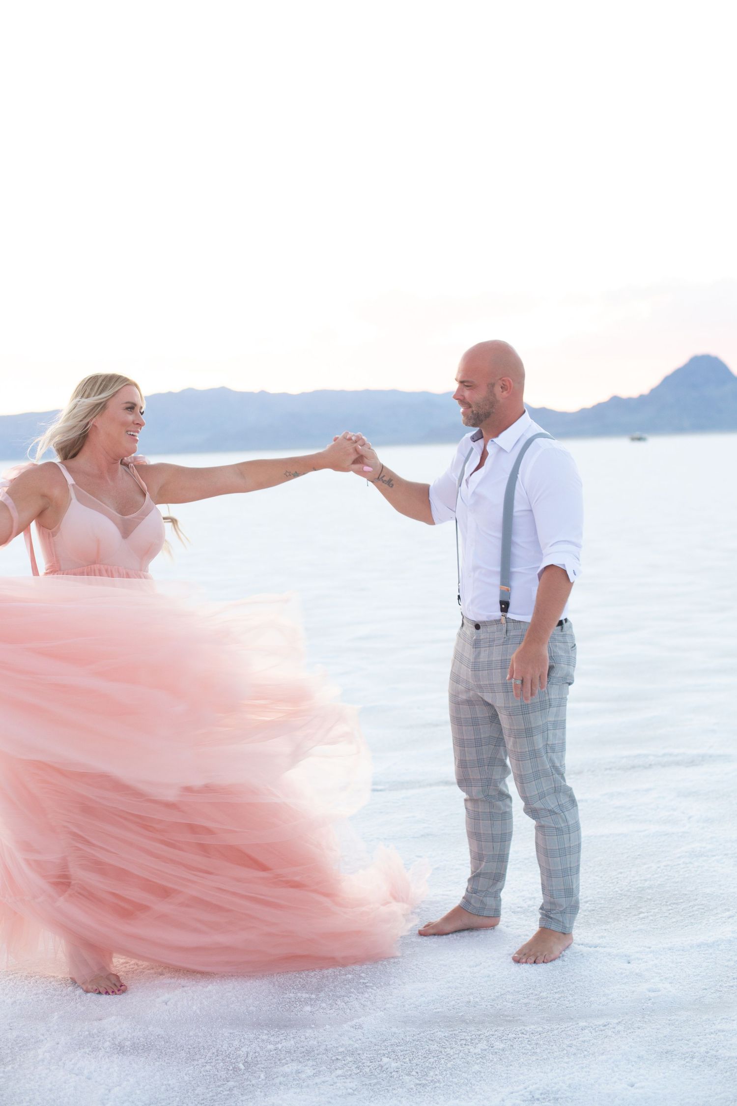 Couple dances playfully on white salt flats, bride wearing flowing pink dress.