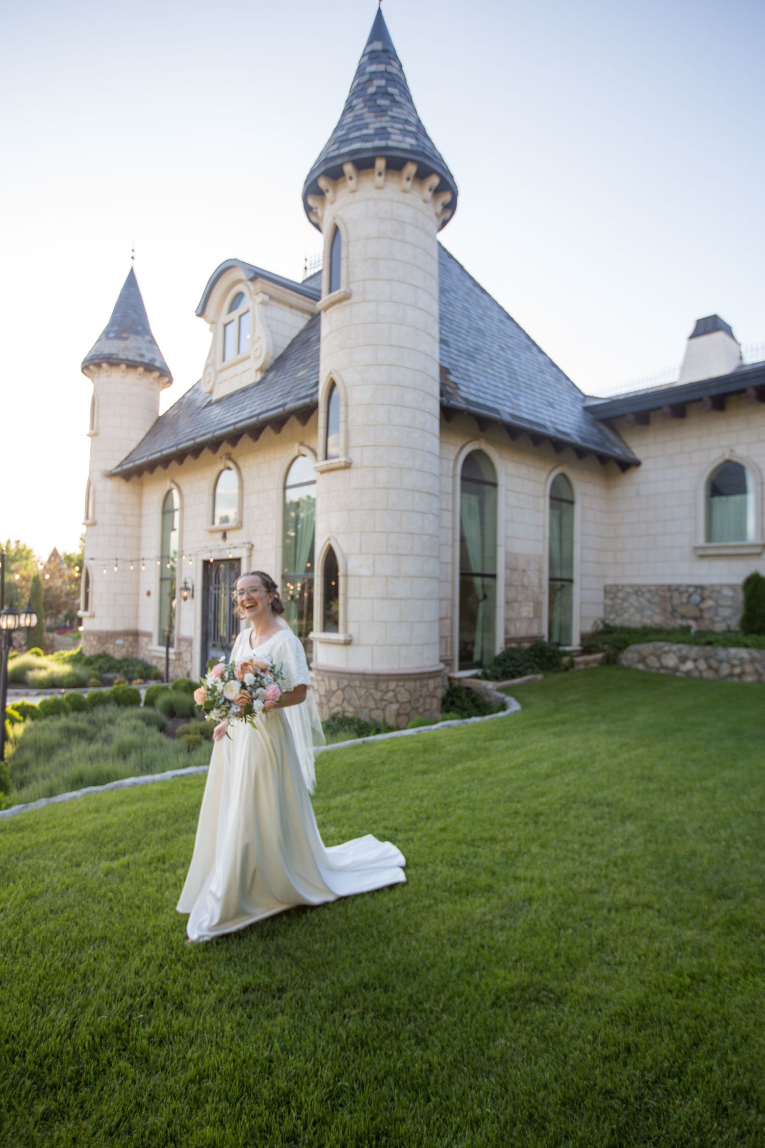 Elegant white chapel with spires provides backdrop for bridal portrait on green lawn at sunset.