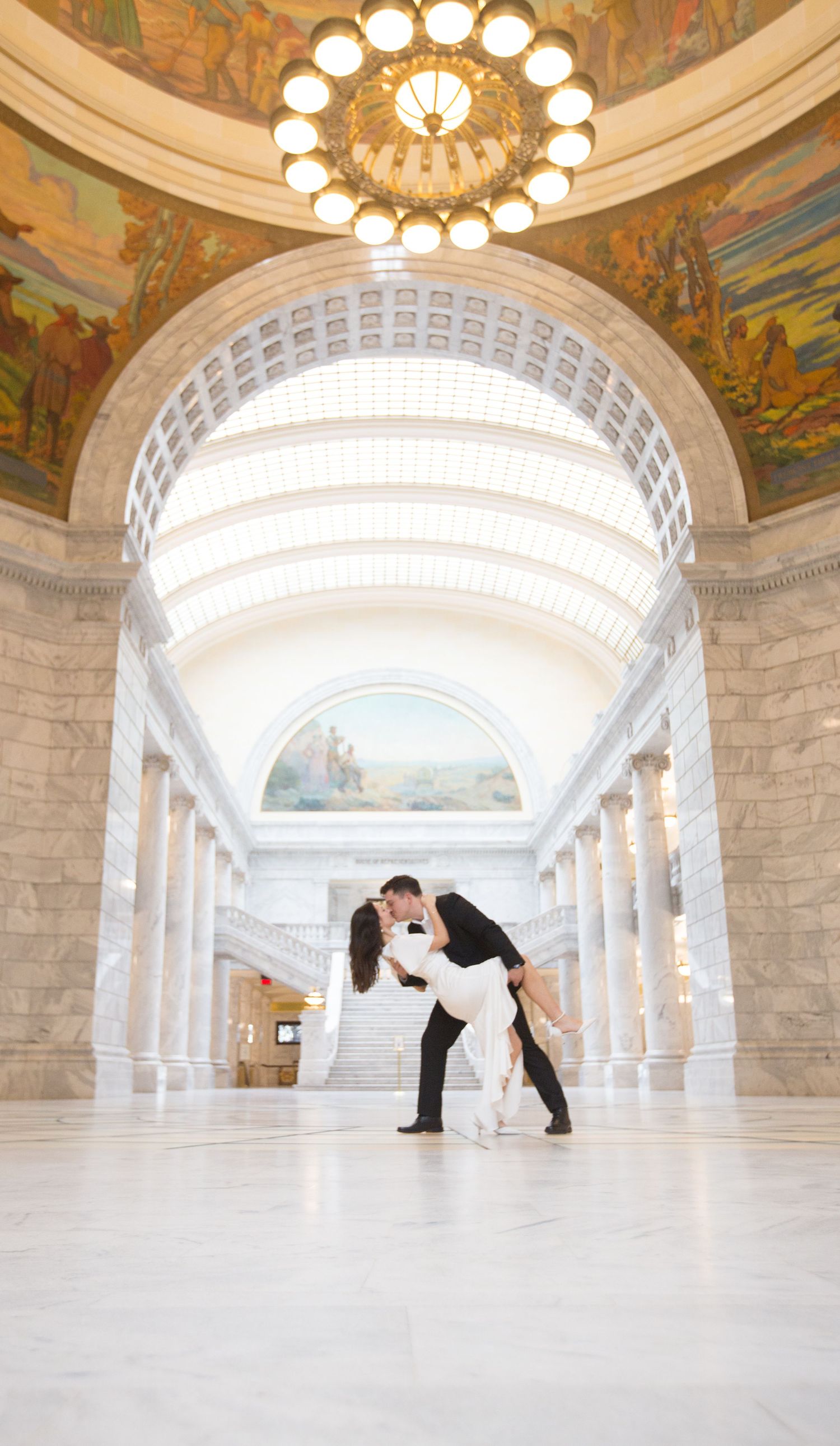 A romantic dip pose in an elegant marble hallway with ornate ceiling and grand lighting.