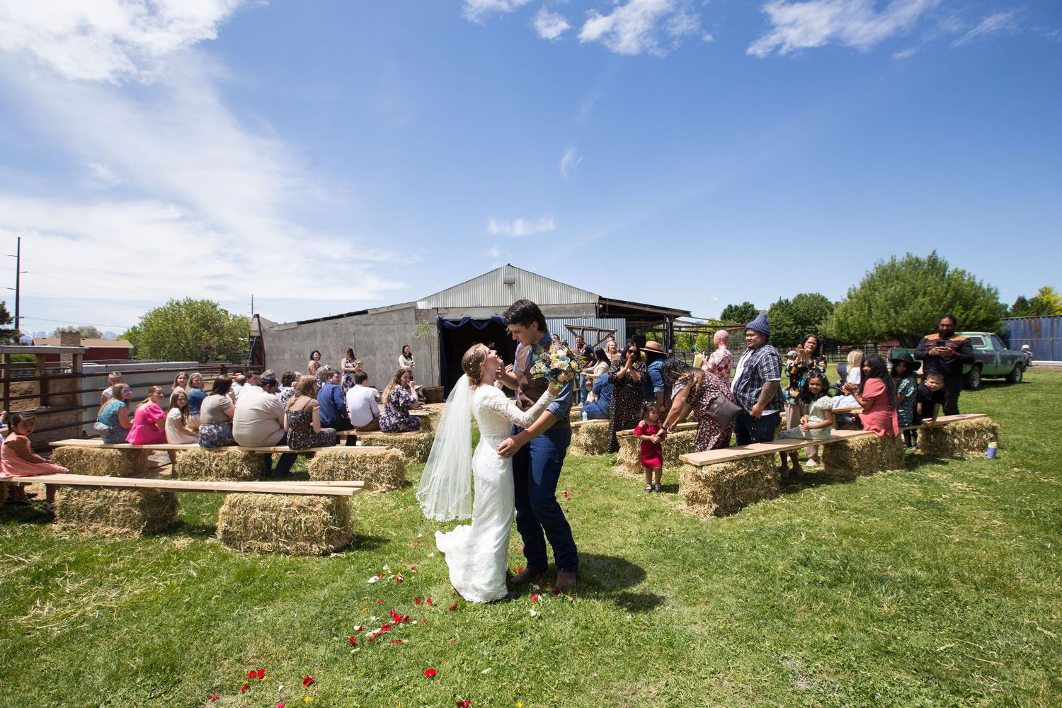 Guests seated on hay bales at an outdoor rustic wedding venue with blue skies overhead.