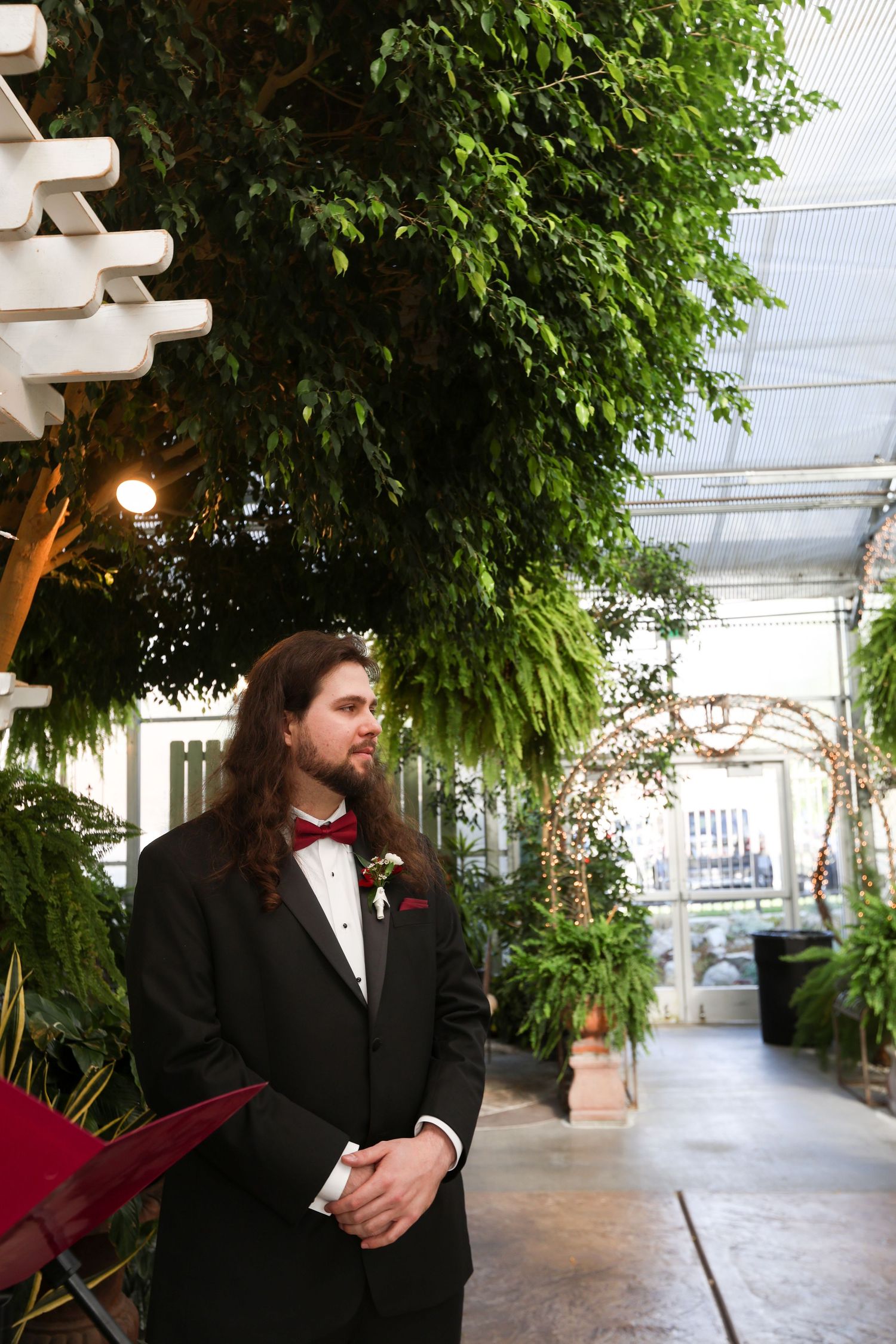 A formal groom in black tuxedo and bowtie waiting in a greenhouse venue.