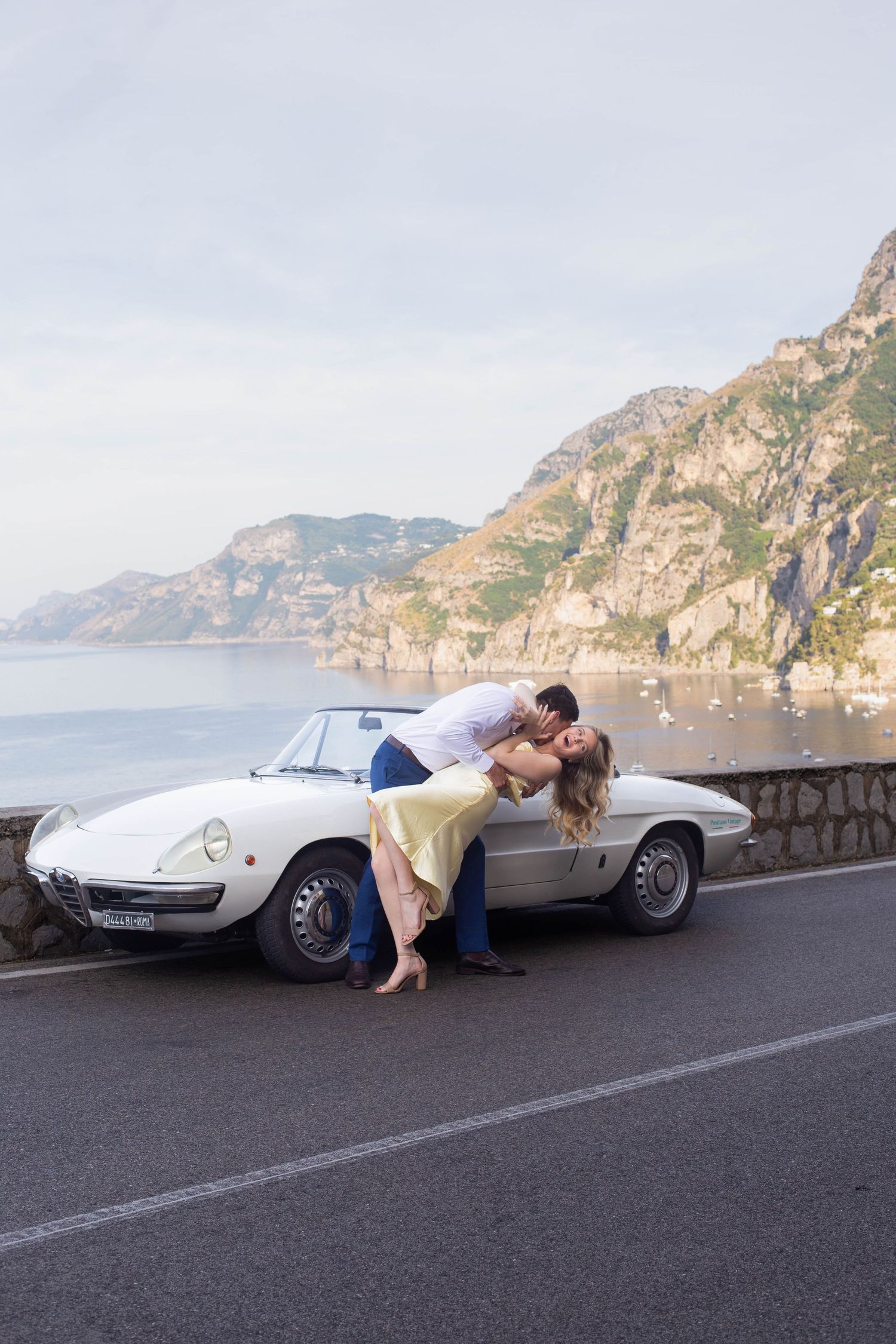 Romantic couple sharing a kiss leaning against a vintage white convertible on the scenic Amalfi Coast road.