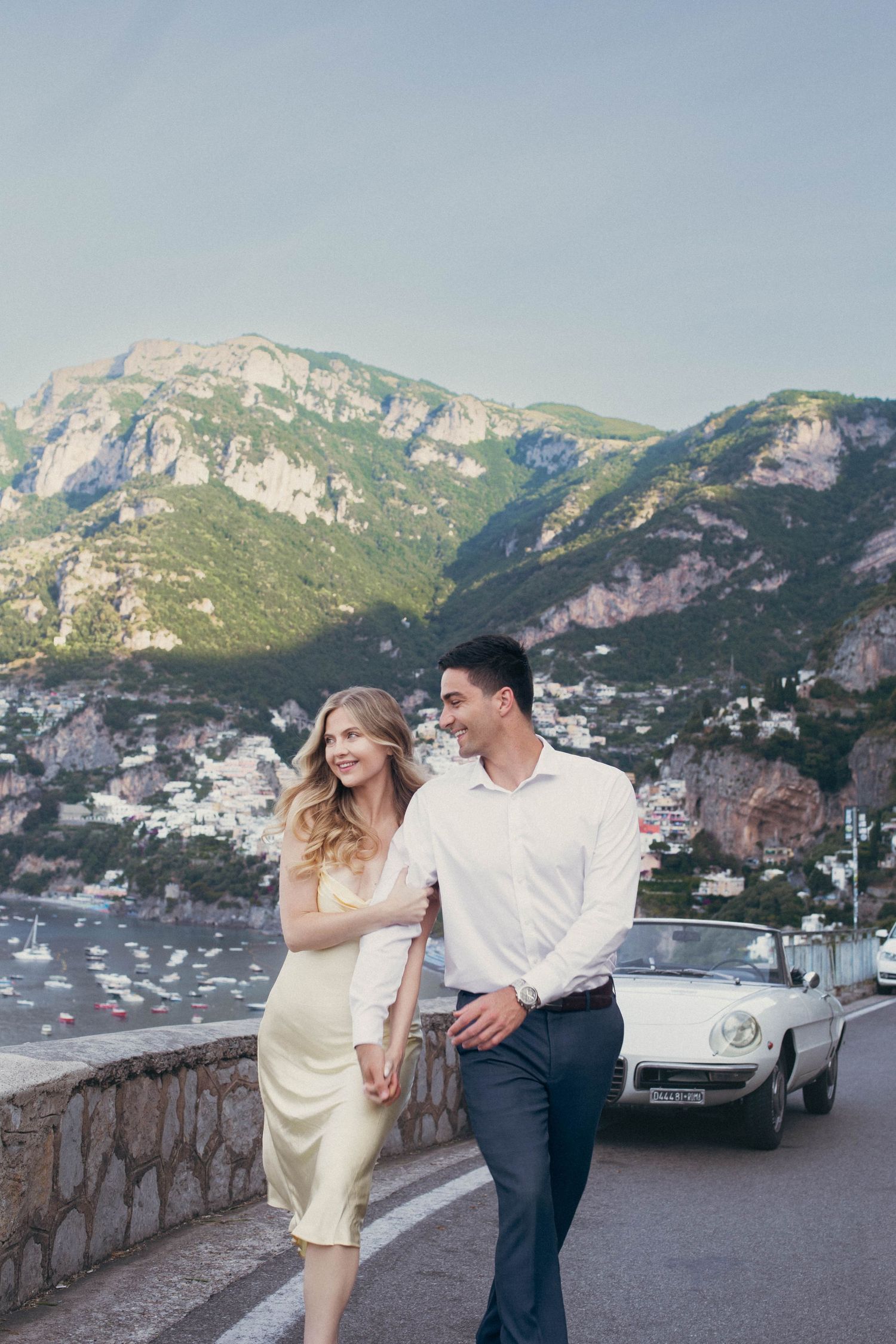 Smiling couple strolling along scenic Positano coastal road beside a classic white convertible sports car.