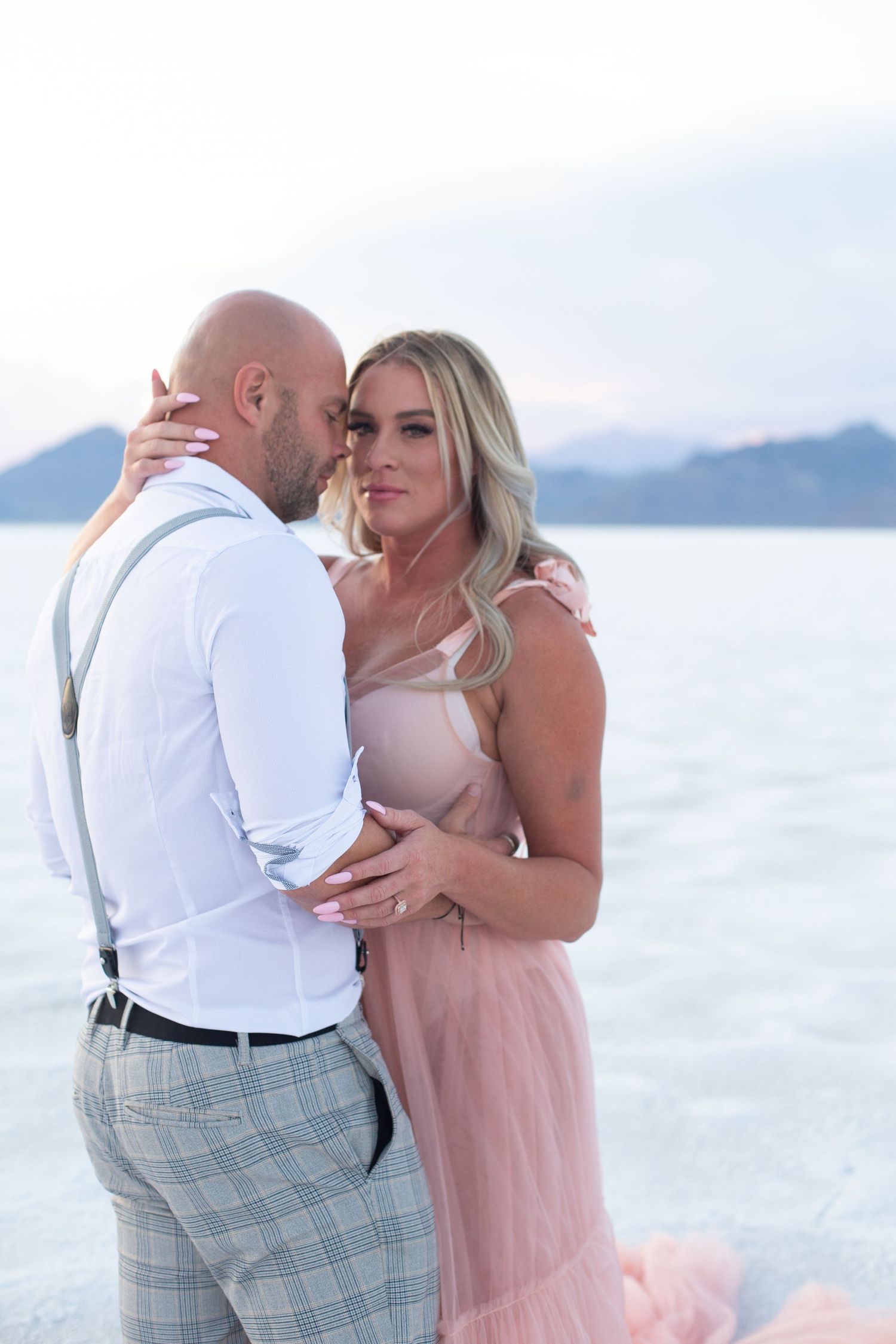 A romantic couple embraces on a salt flat at sunset, with the woman wearing a flowing pink gown.