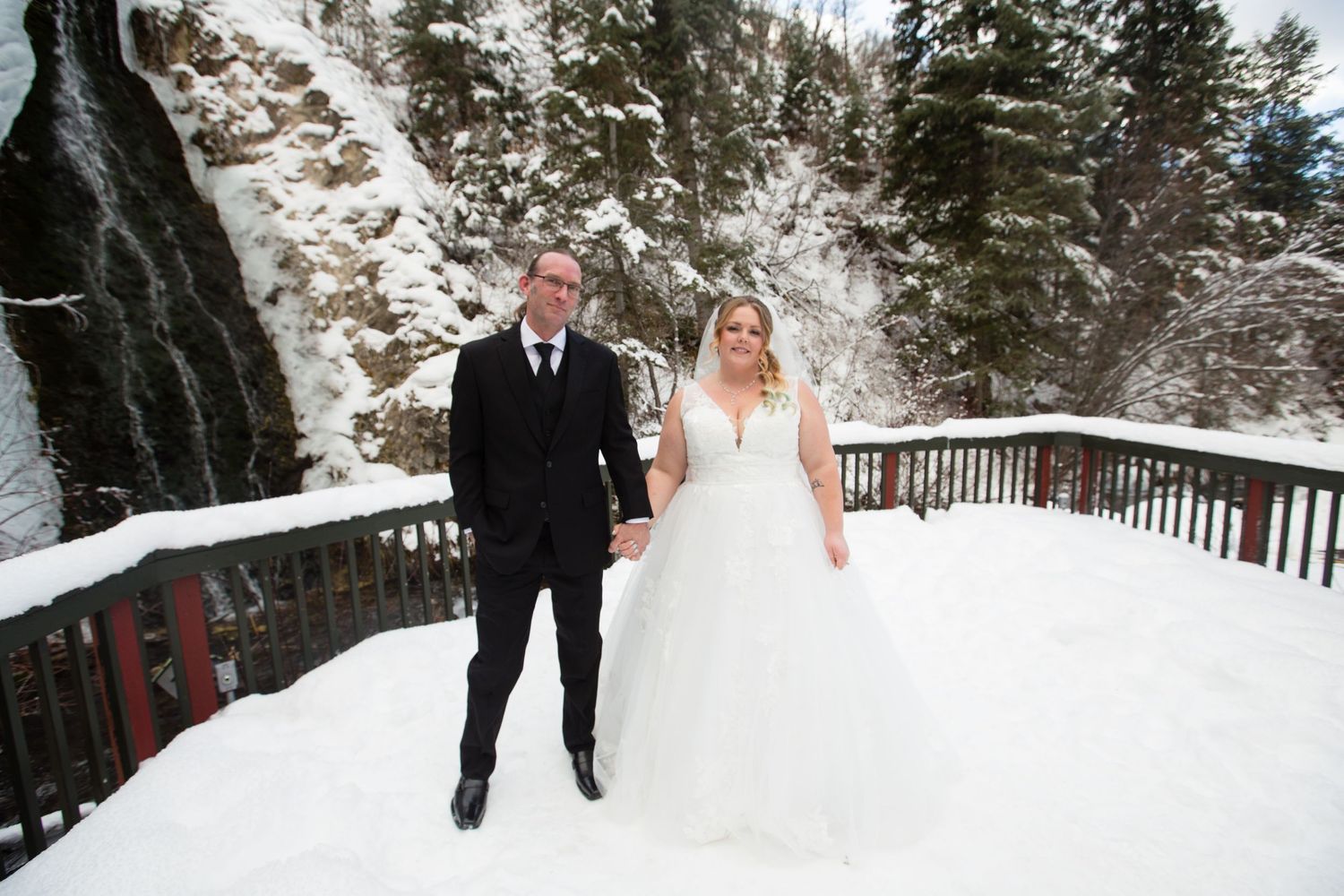 Couple walking along a snow-covered wooden deck surrounded by evergreen trees.