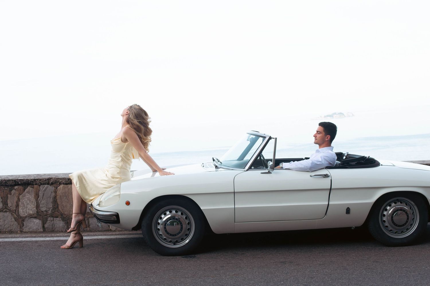 Elegant couple in a white convertible sports car parked by the sea, woman in yellow dress leaning on the hood.