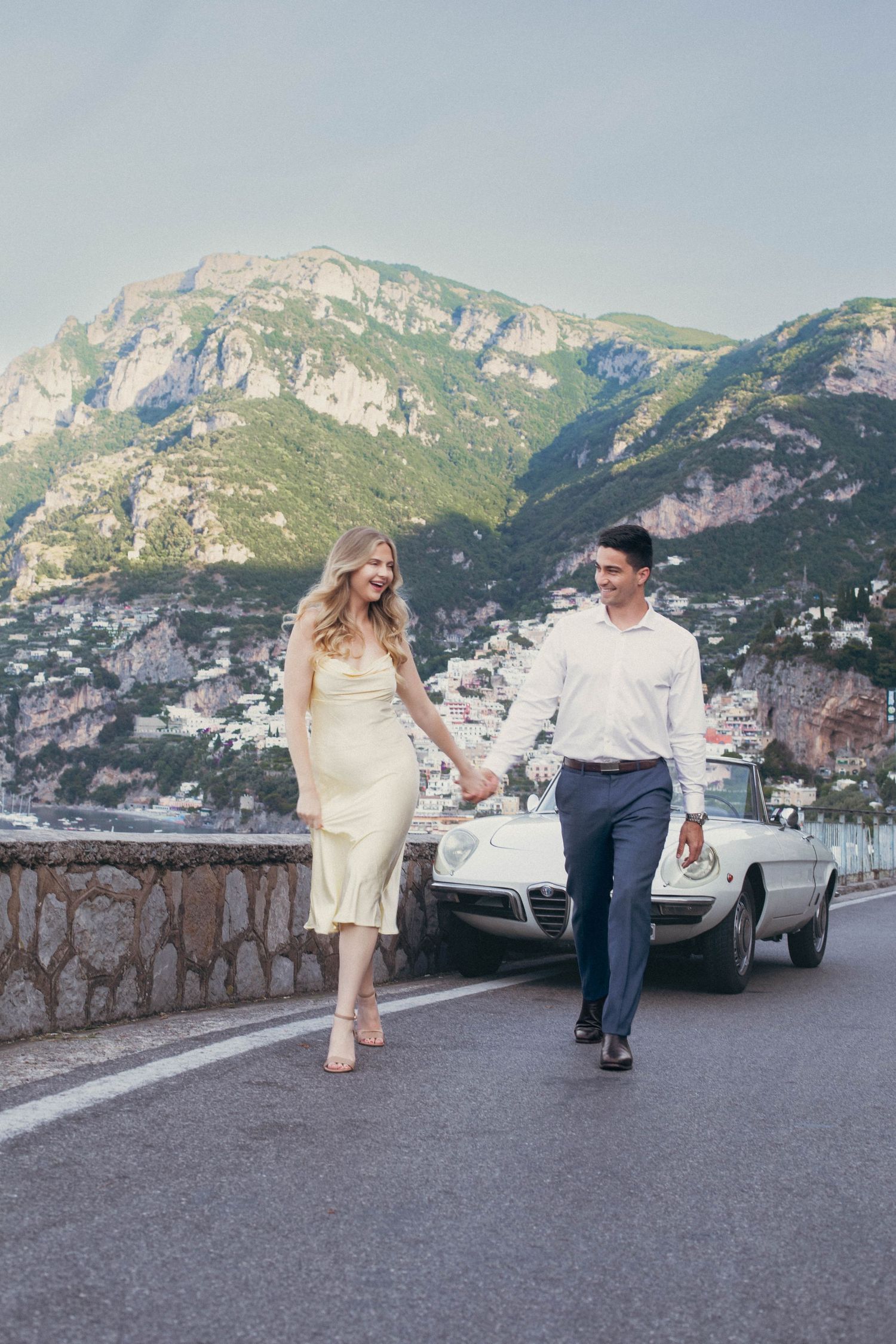 Couple holding hands walking on Amalfi Coast road with vintage white car and cliffside village backdrop.