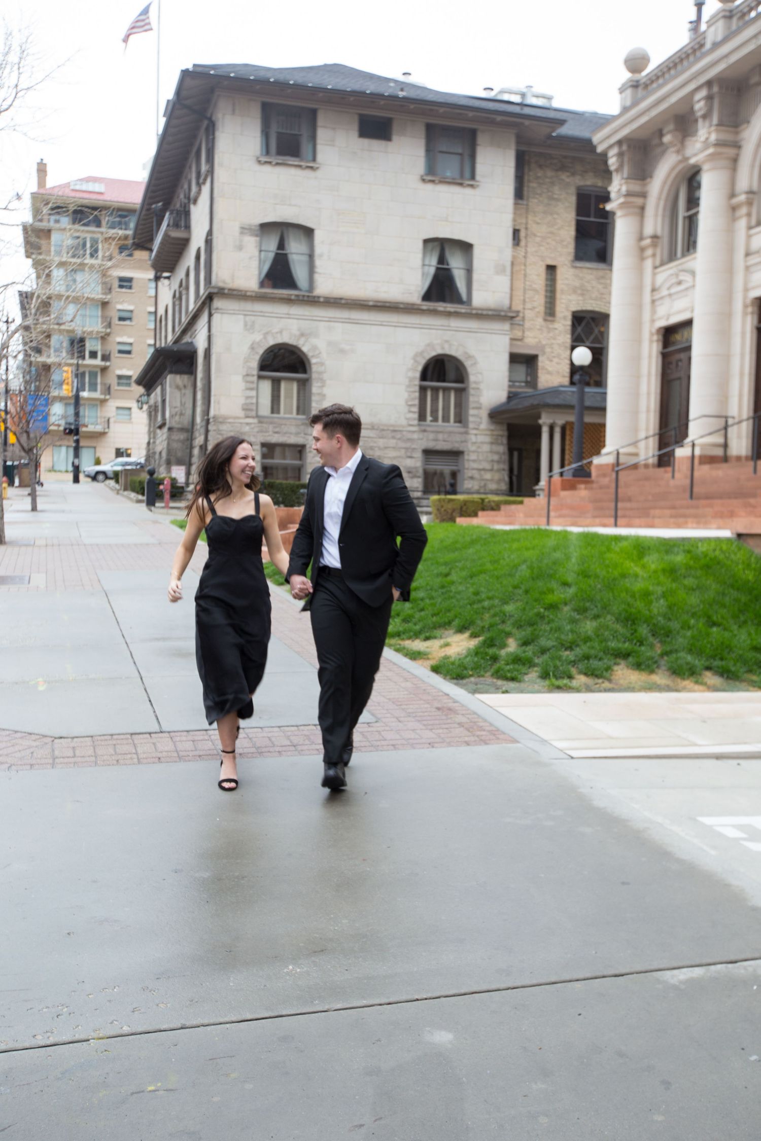 A stylish couple in black formal attire walks hand-in-hand laughing along an urban sidewalk.