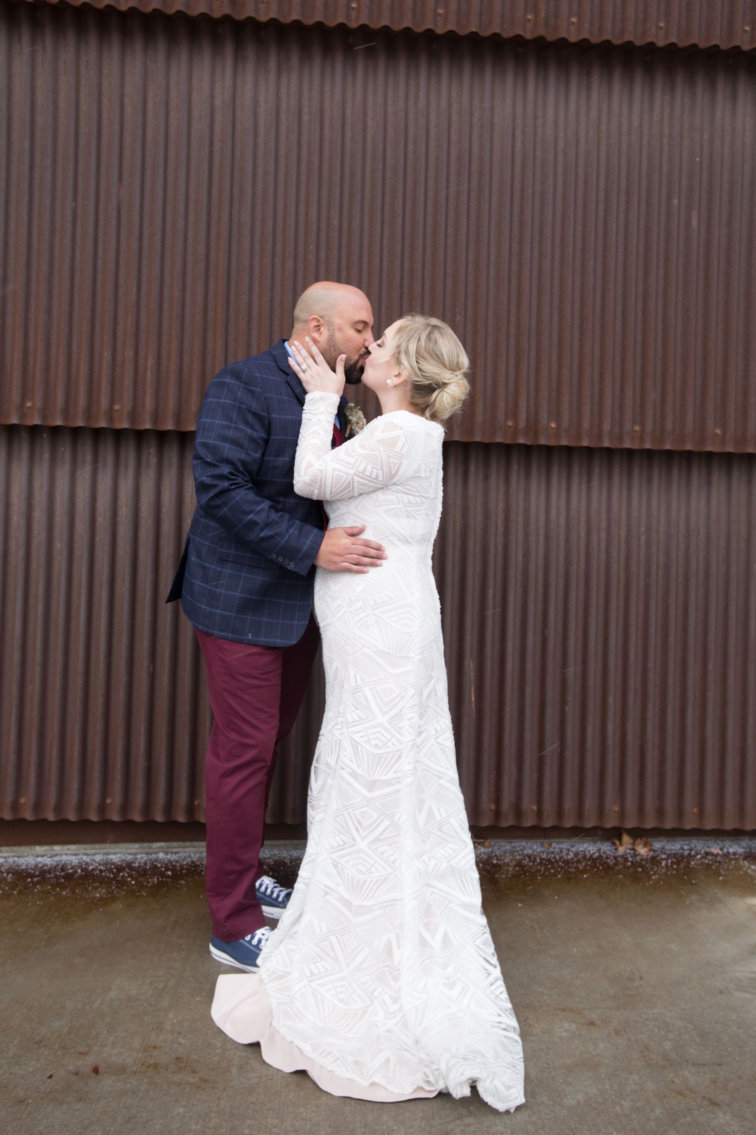 Bride and groom share a romantic kiss in front of a rustic corrugated metal wall, bride in elegant white lace gown.