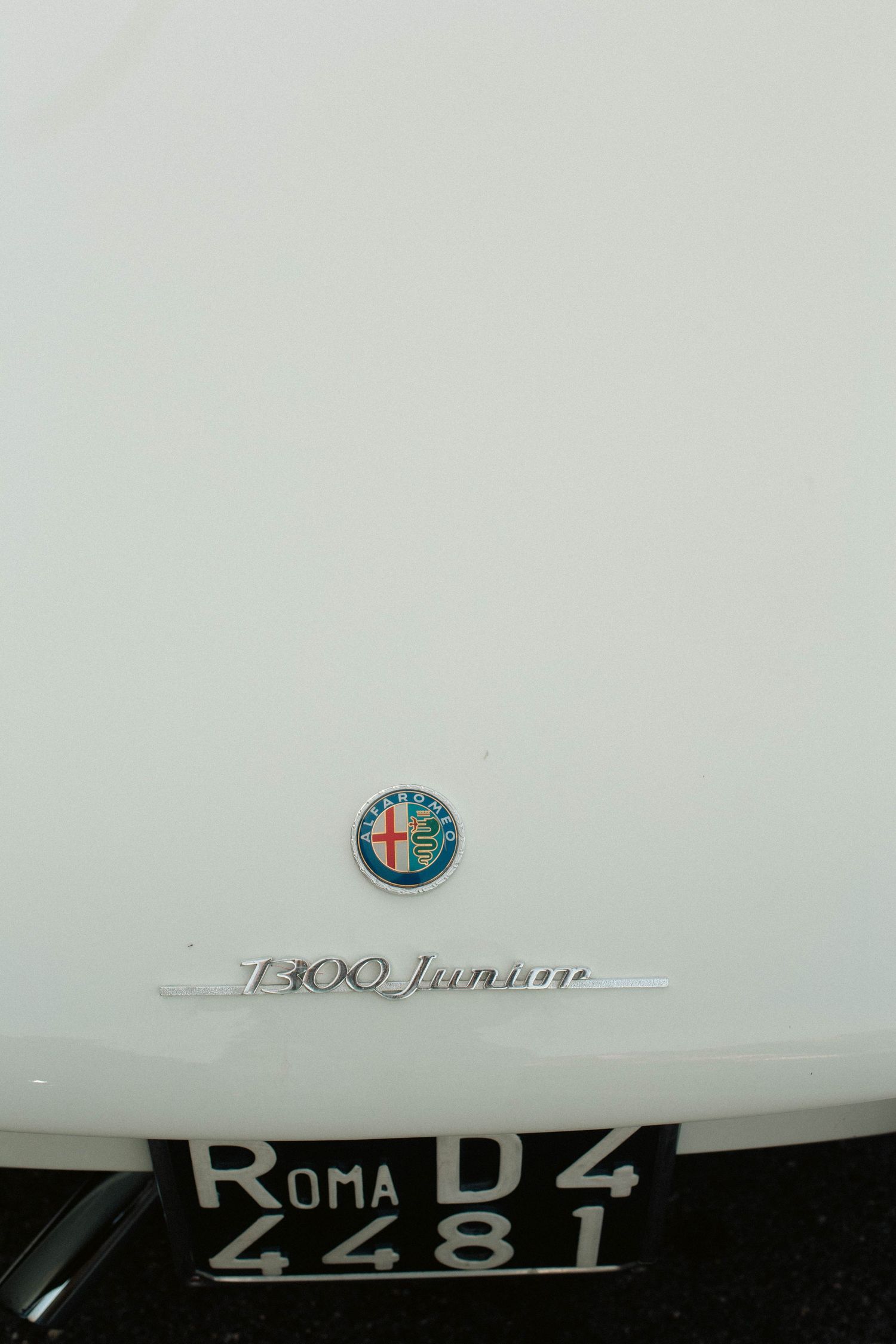 Close-up of Alfa Romeo 1300 Junior badge and Roma license plate on white vintage car.