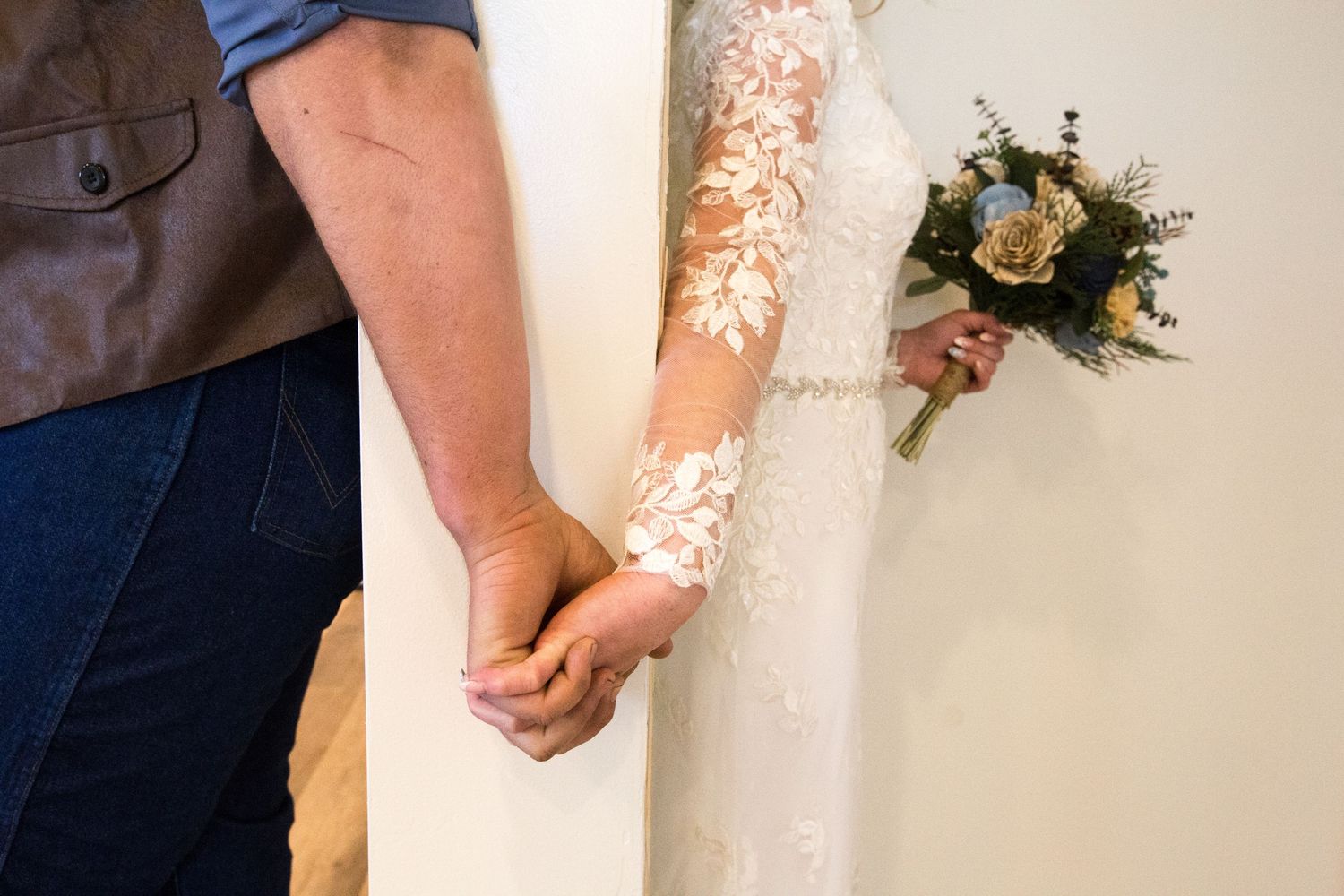 Bride and groom holding hands, bride wearing lace wedding gown holding blue floral bouquet.