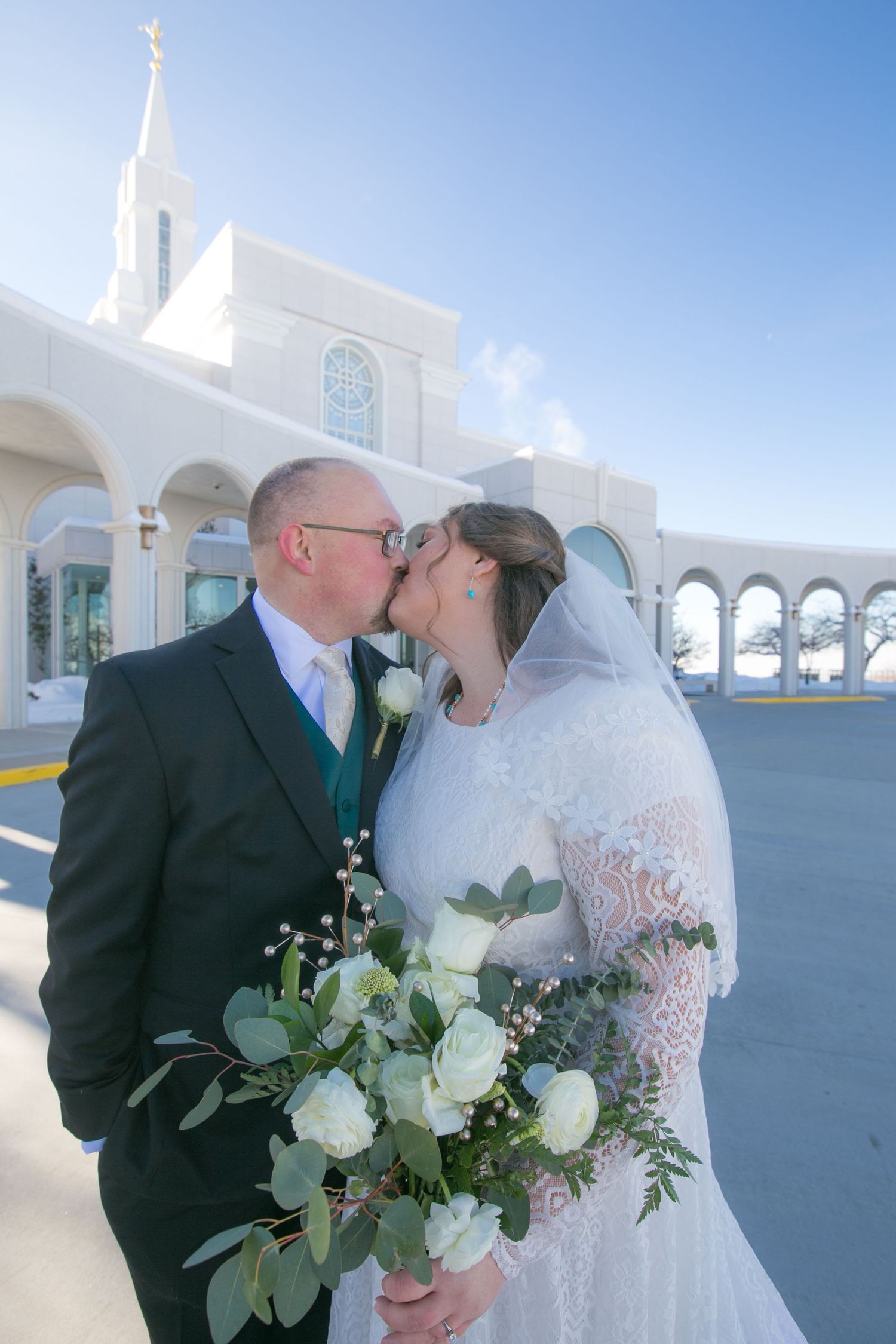 Newlywed couple kissing in front of a white LDS temple, bride holding white rose bouquet with eucalyptus on sunny day.