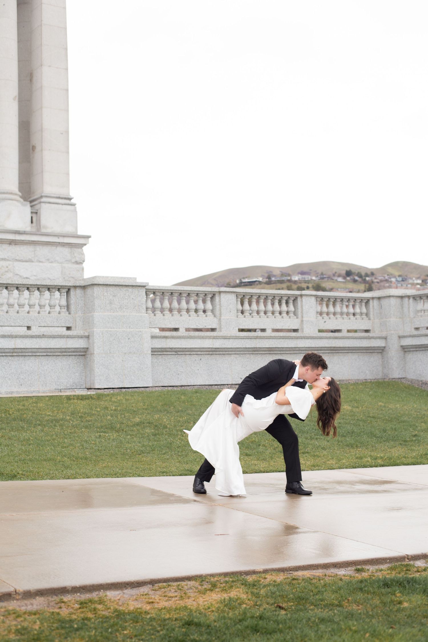 A groom dips his bride for a romantic kiss outside a grand white stone building on their wedding day.