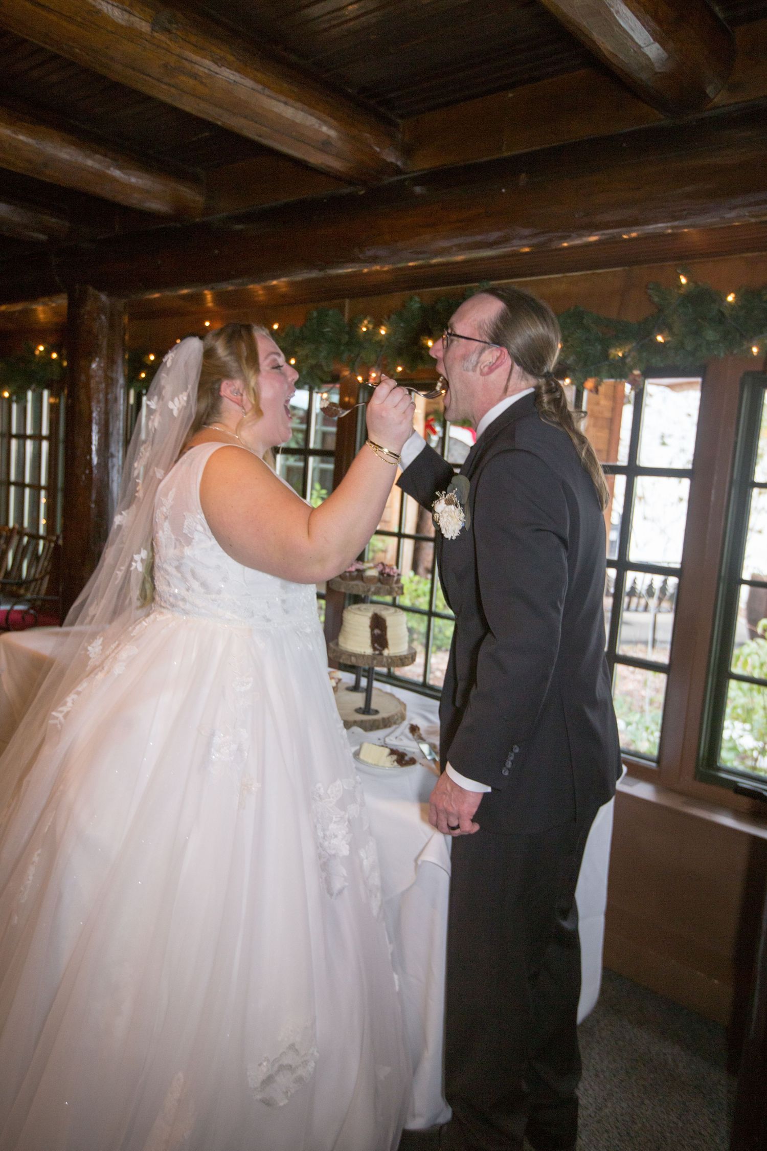 Bride feeds groom wedding cake during reception inside rustic log cabin venue decorated with garland and fairy lights.