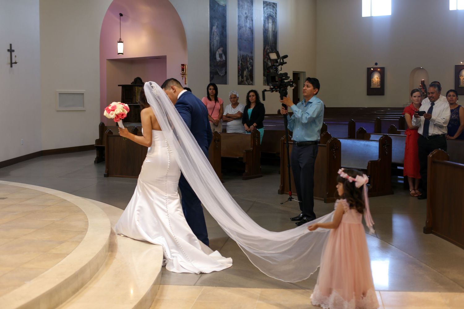 A couple shares their first dance in a church while guests look on and a small flower girl watches in a pink dress.