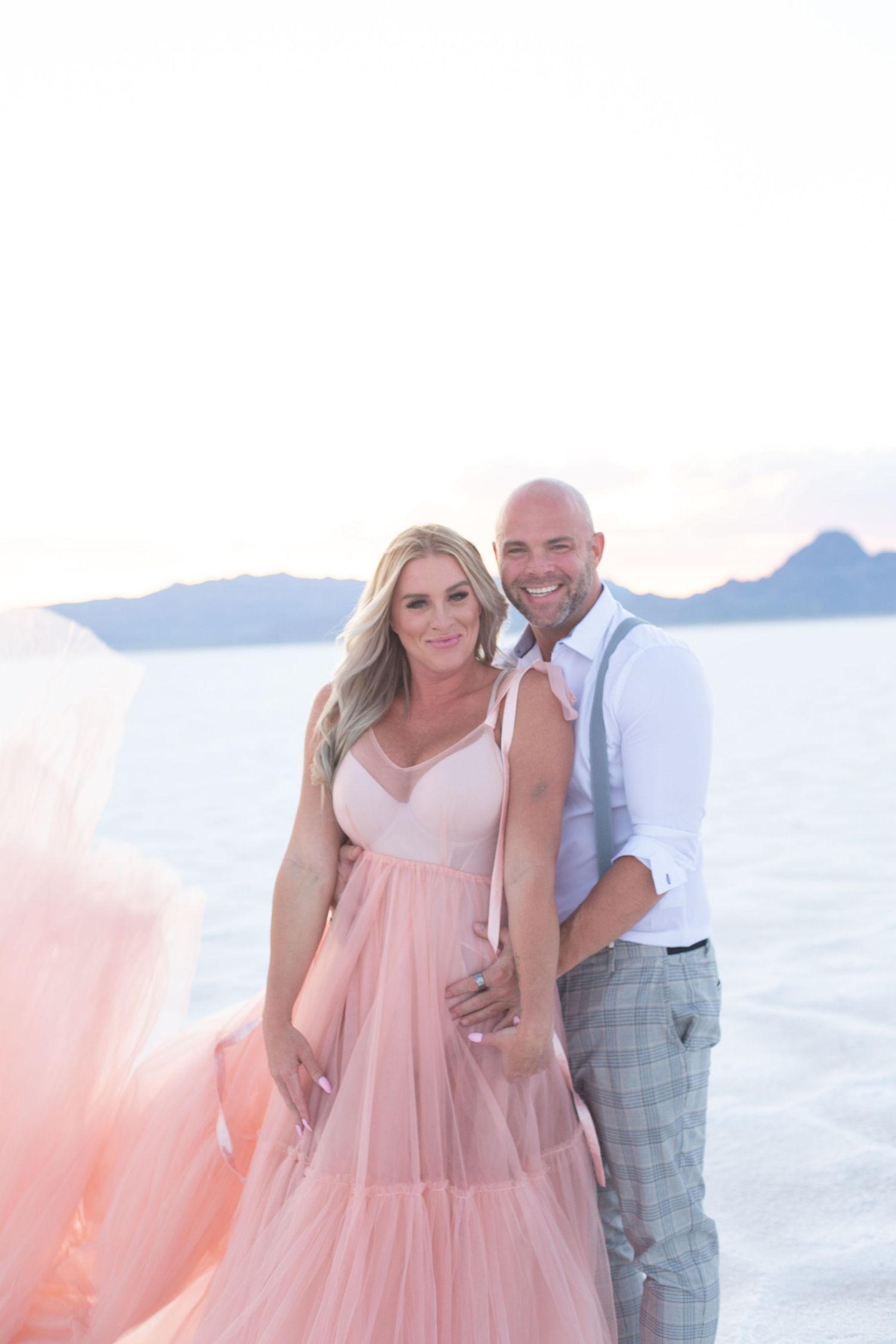 Couple poses together against ocean backdrop wearing pink dress and plaid pants.