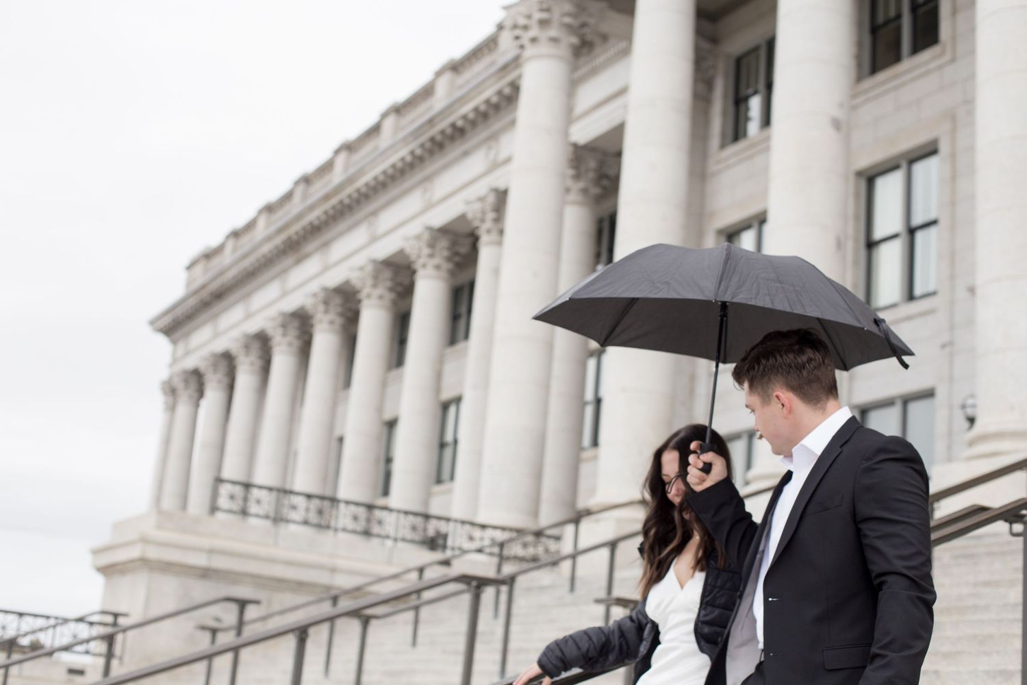 Couple poses with umbrellas outside a grand historic building with columns on a rainy day.