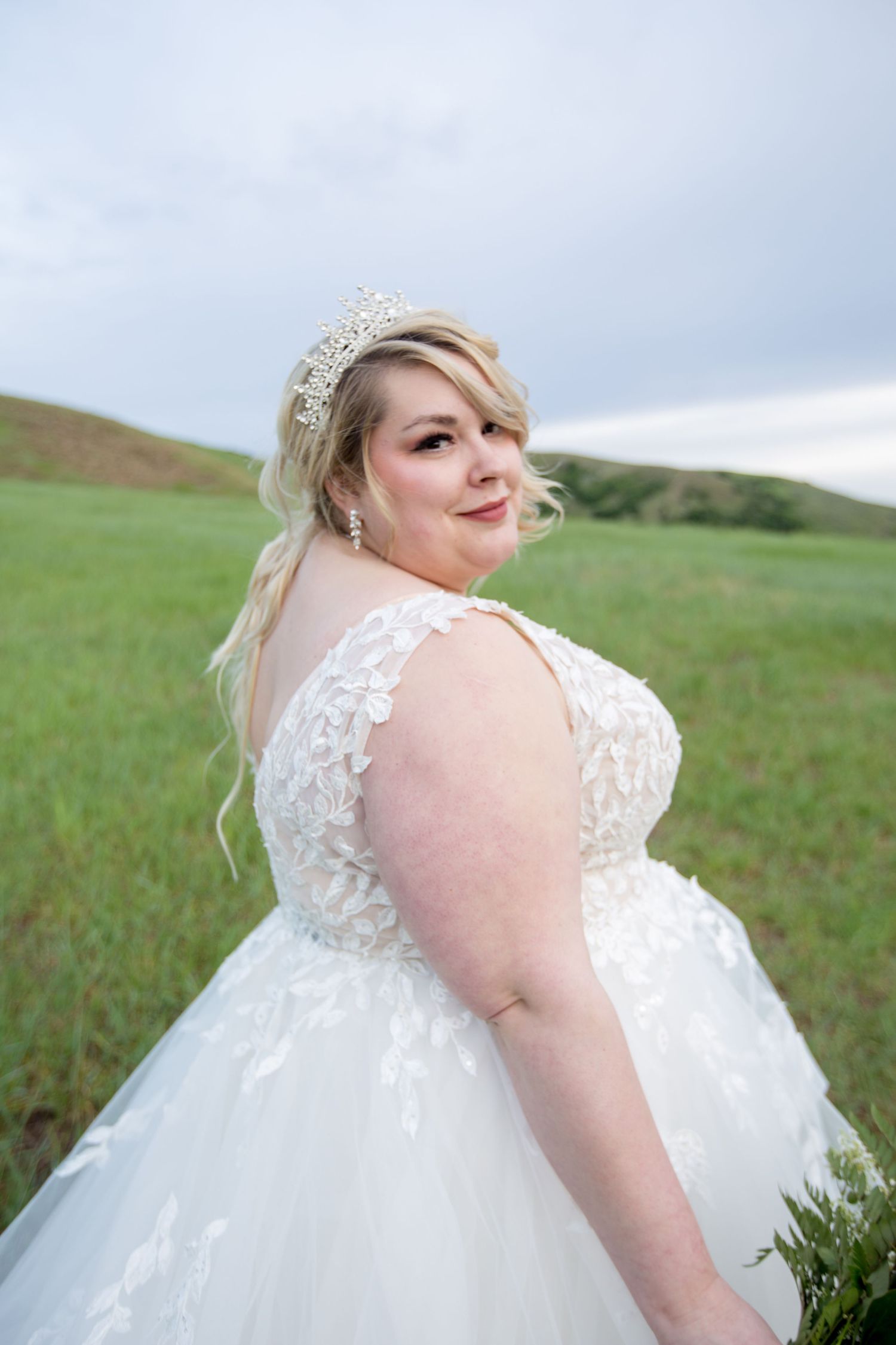 Elegant lace detail on white wedding dress shown against green hillside background.