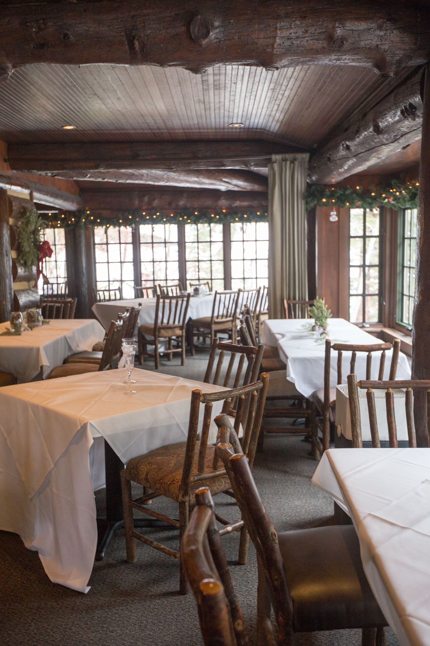 Rustic log cabin restaurant dining room with white tablecloths, wooden chairs, and festive Christmas garland decorating the windows.