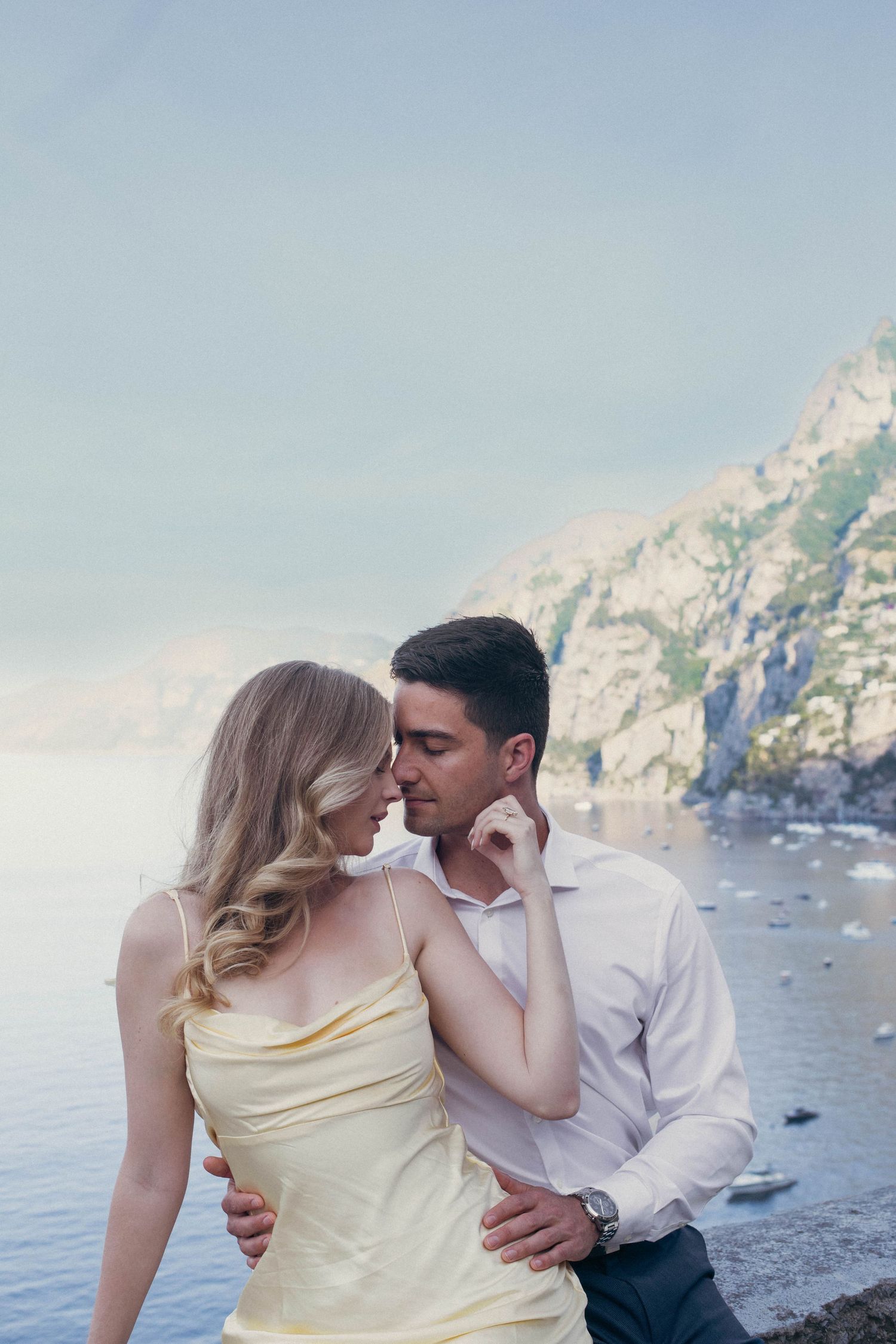 Elegant couple in intimate embrace near rocky Amalfi Coast shoreline with calm sea and cliffs behind.