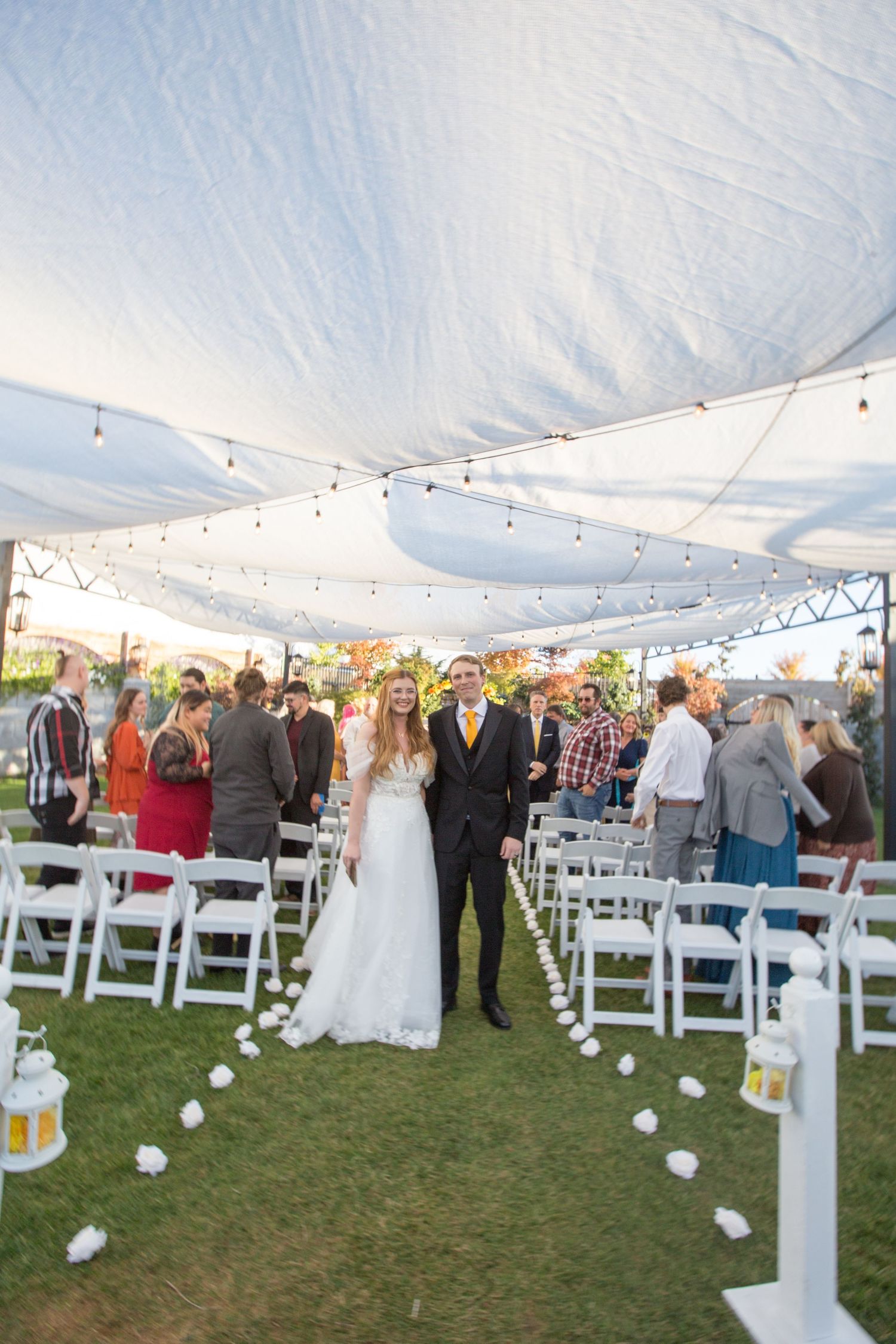 Outdoor wedding ceremony setup with white tent, string lights, and white folding chairs on grass.