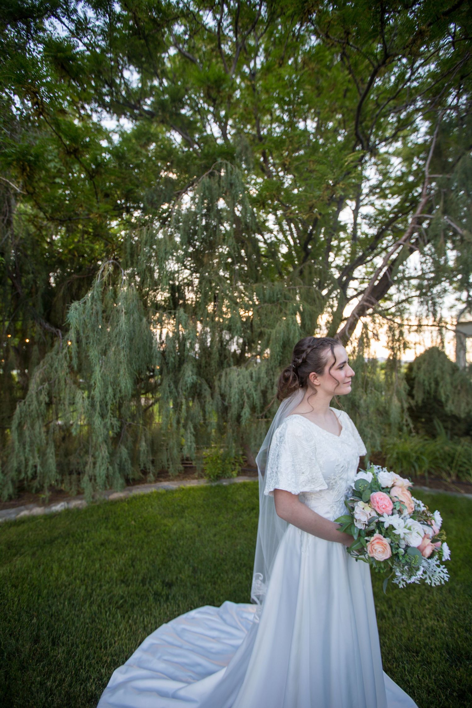 A white wedding dress with train spreads elegantly across green grass in a garden setting.