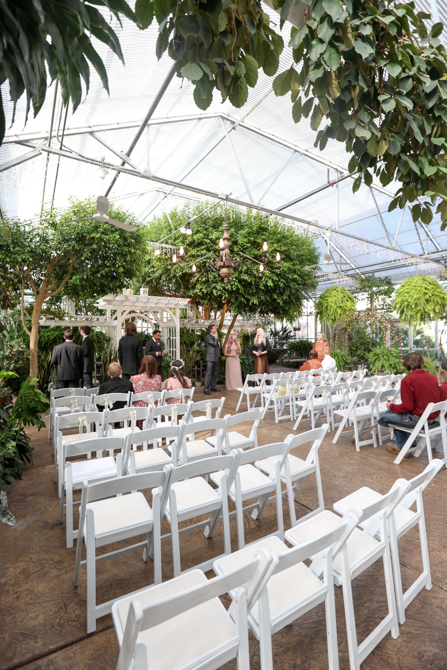 White folding chairs arranged in rows inside a greenhouse wedding venue with lush greenery overhead.