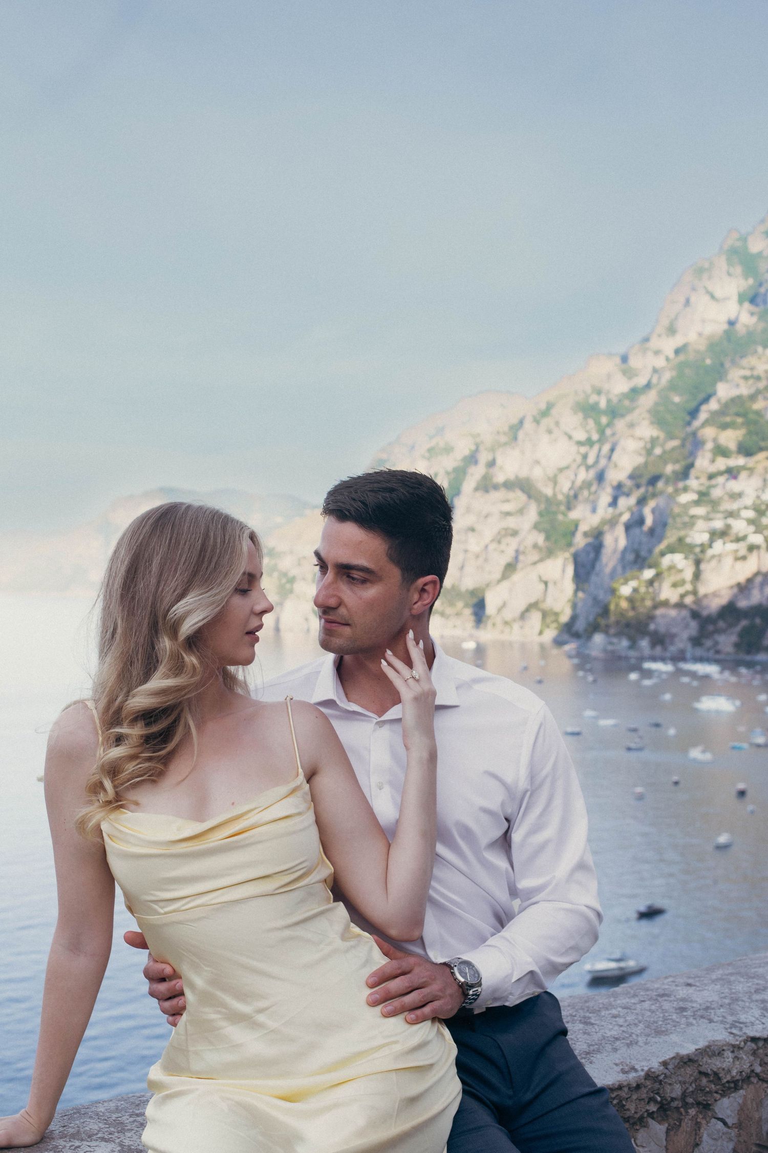 Romantic couple embracing by stone wall with breathtaking Amalfi Coast sea and mountain backdrop at dusk.