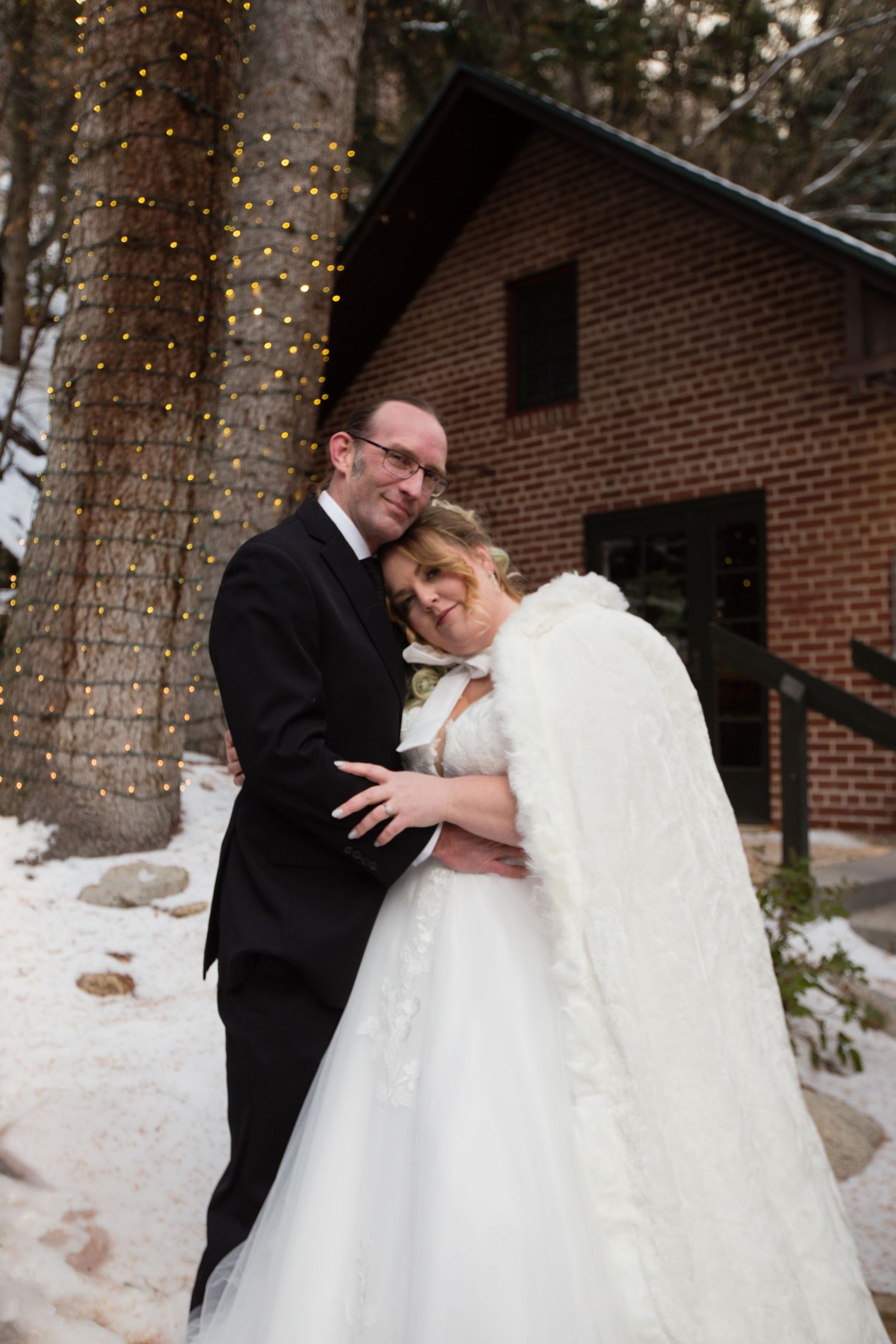 Wedding couple shares romantic moments under trees decorated with twinkling lights in snowy setting.