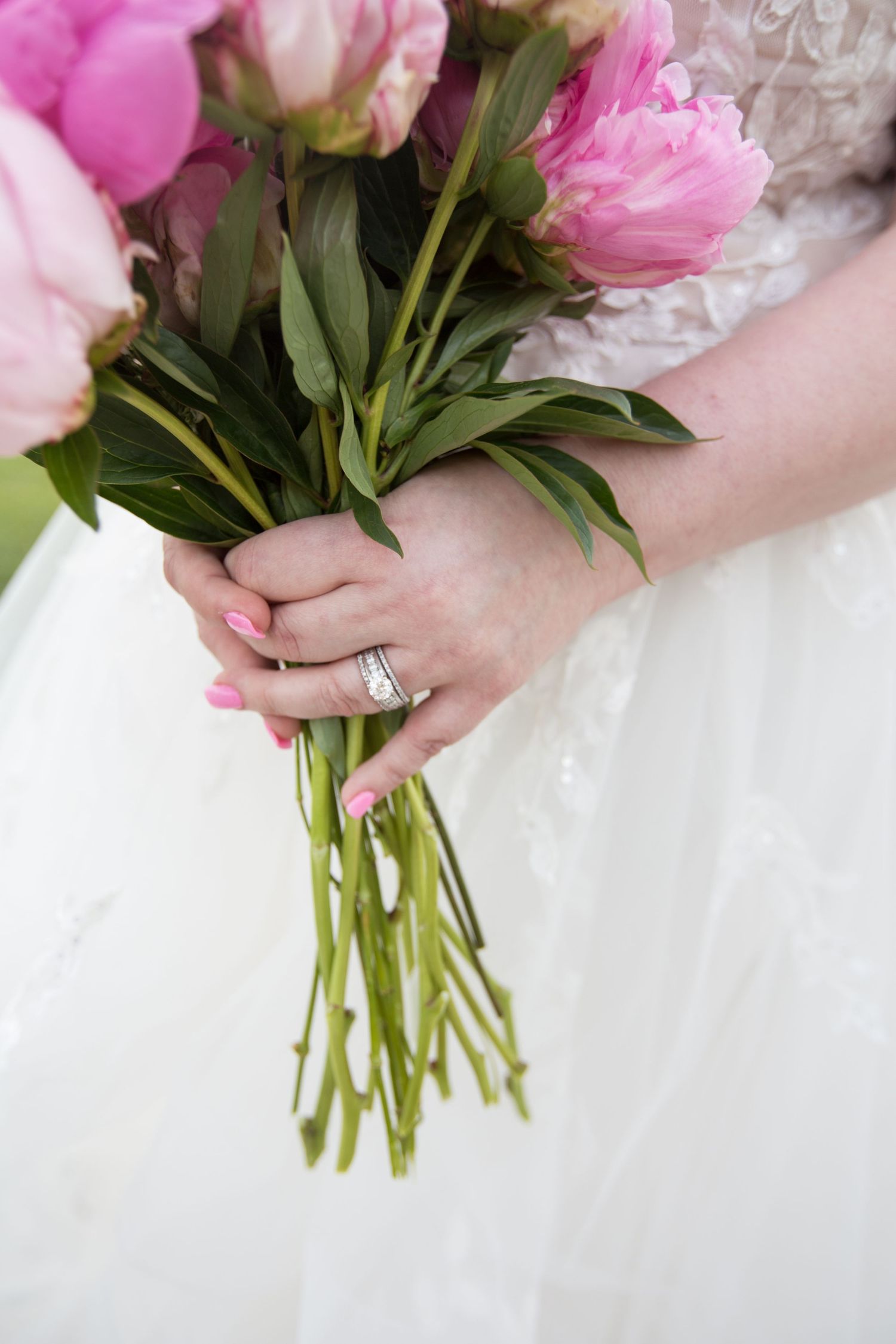 Close-up of hands holding pink peony bouquet with engagement ring visible.