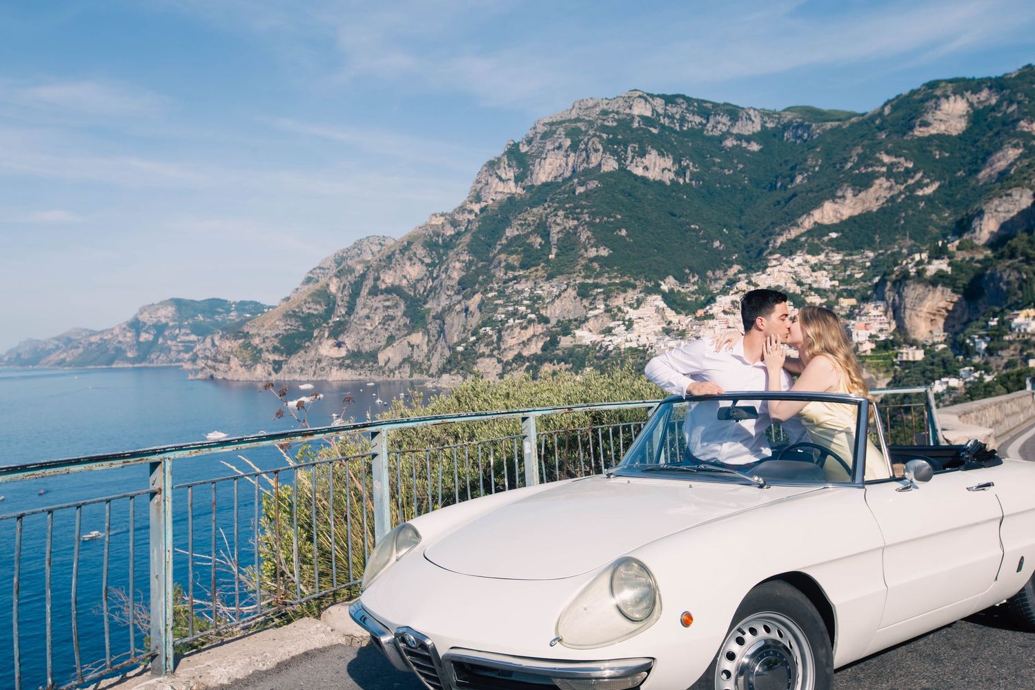 Couple kissing in classic white Alfa Romeo Spider convertible on scenic Amalfi Coast road.